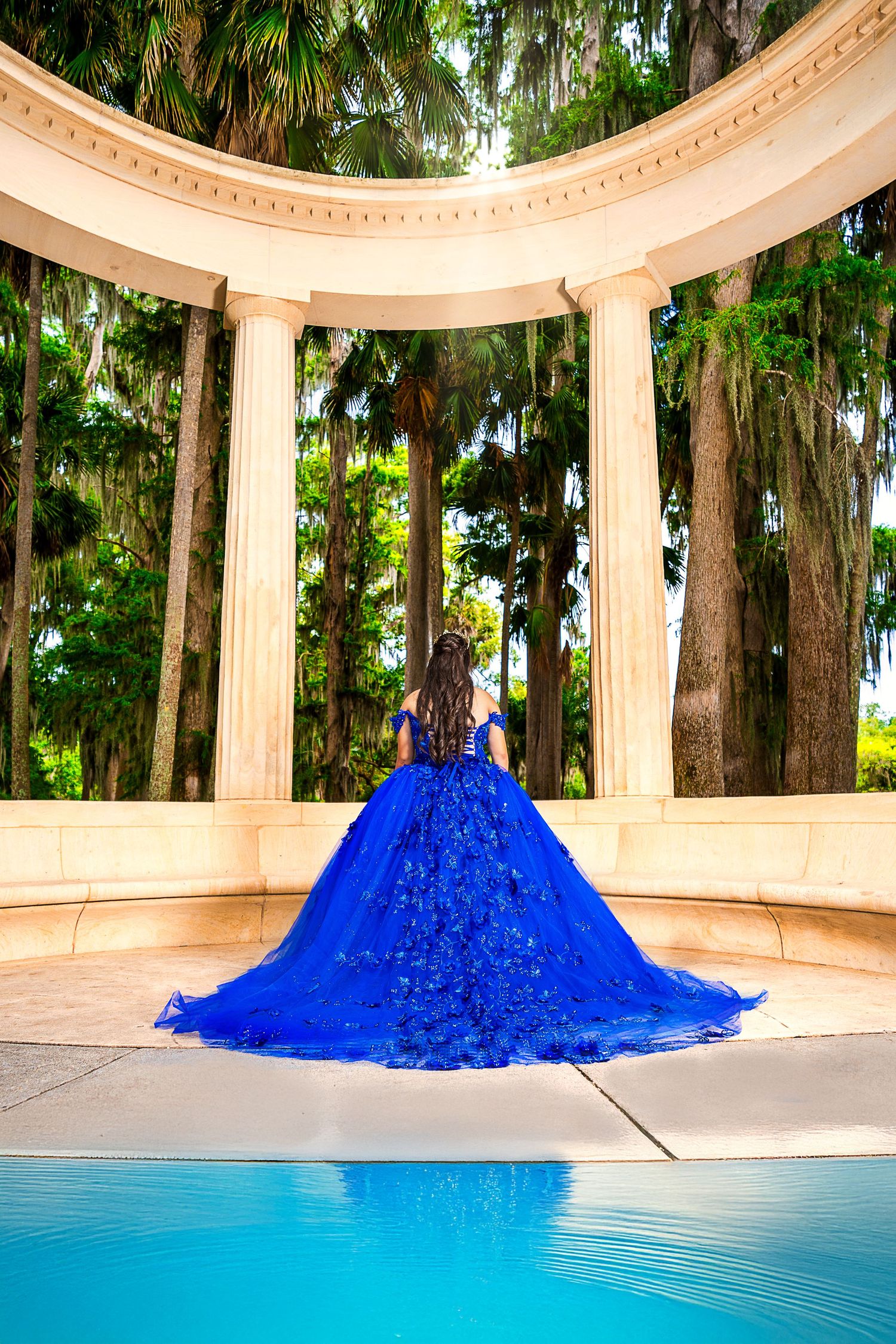 Young woman in royal blue quinceañera ball gown poses by pool at elegant columned rotunda surrounded by palm trees.