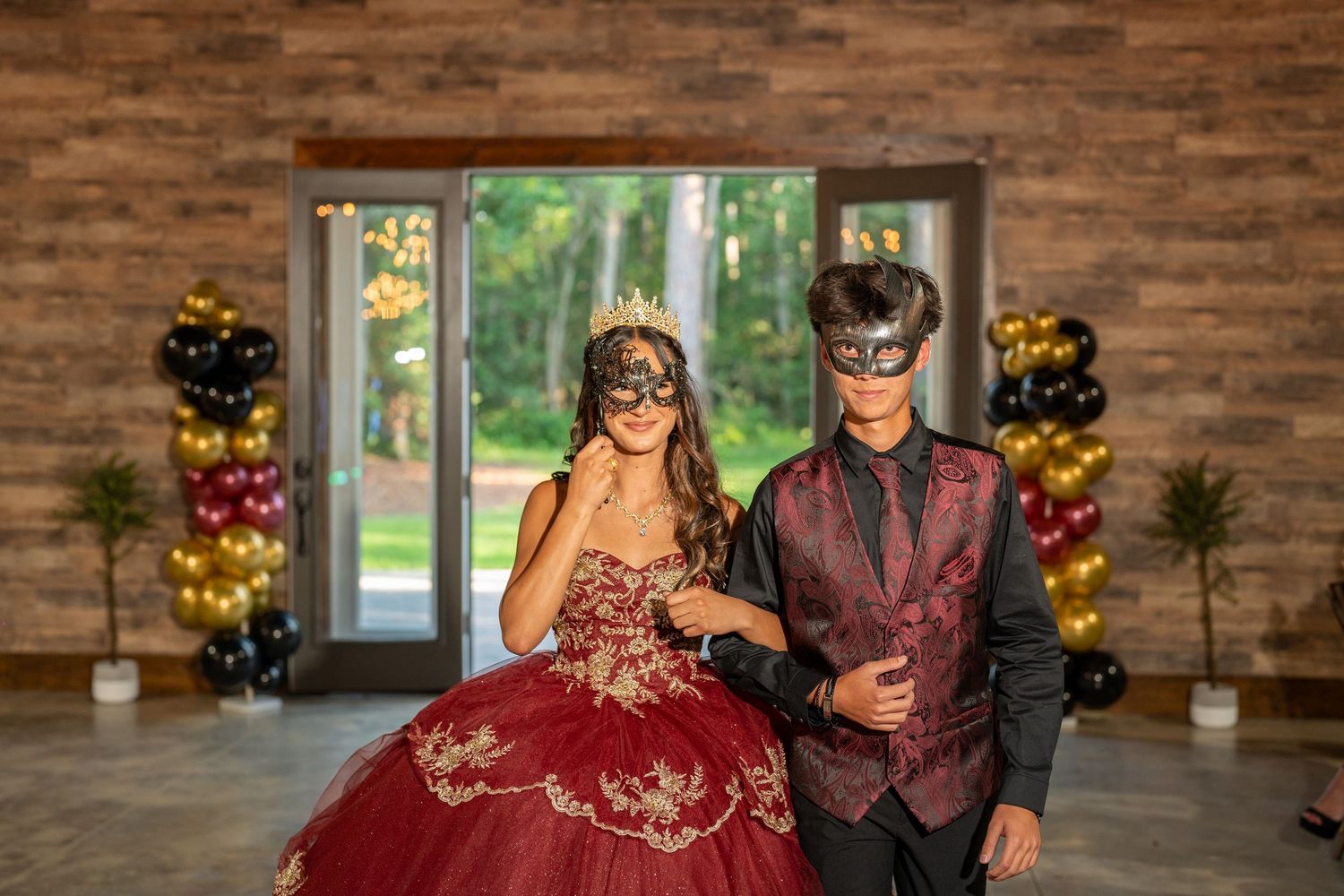 Couple in masquerade masks posing at quinceañera, wearing red ballgown and burgundy suit with gold accents.