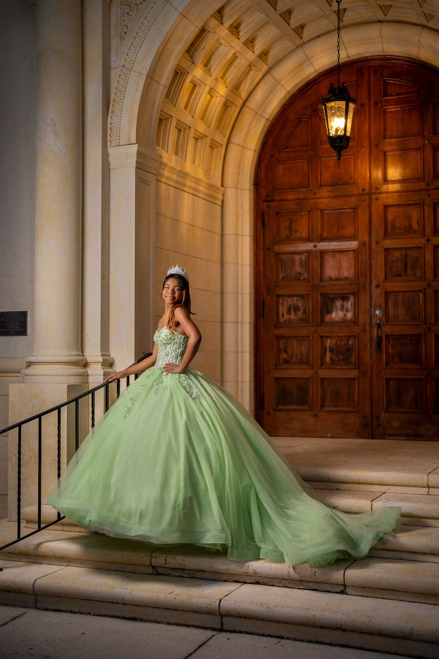 Young woman in green ball gown and tiara poses on steps before ornate wooden doors at quinceañera.