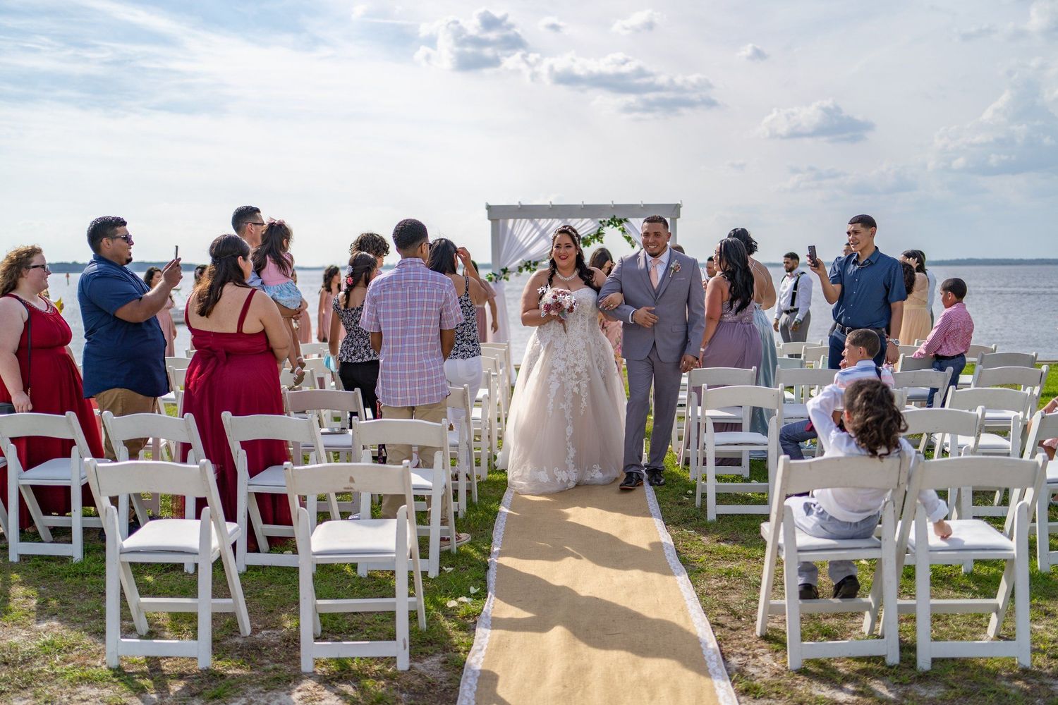 Outdoor beach wedding ceremony with bride and groom walking down aisle lined with white chairs and guests.
