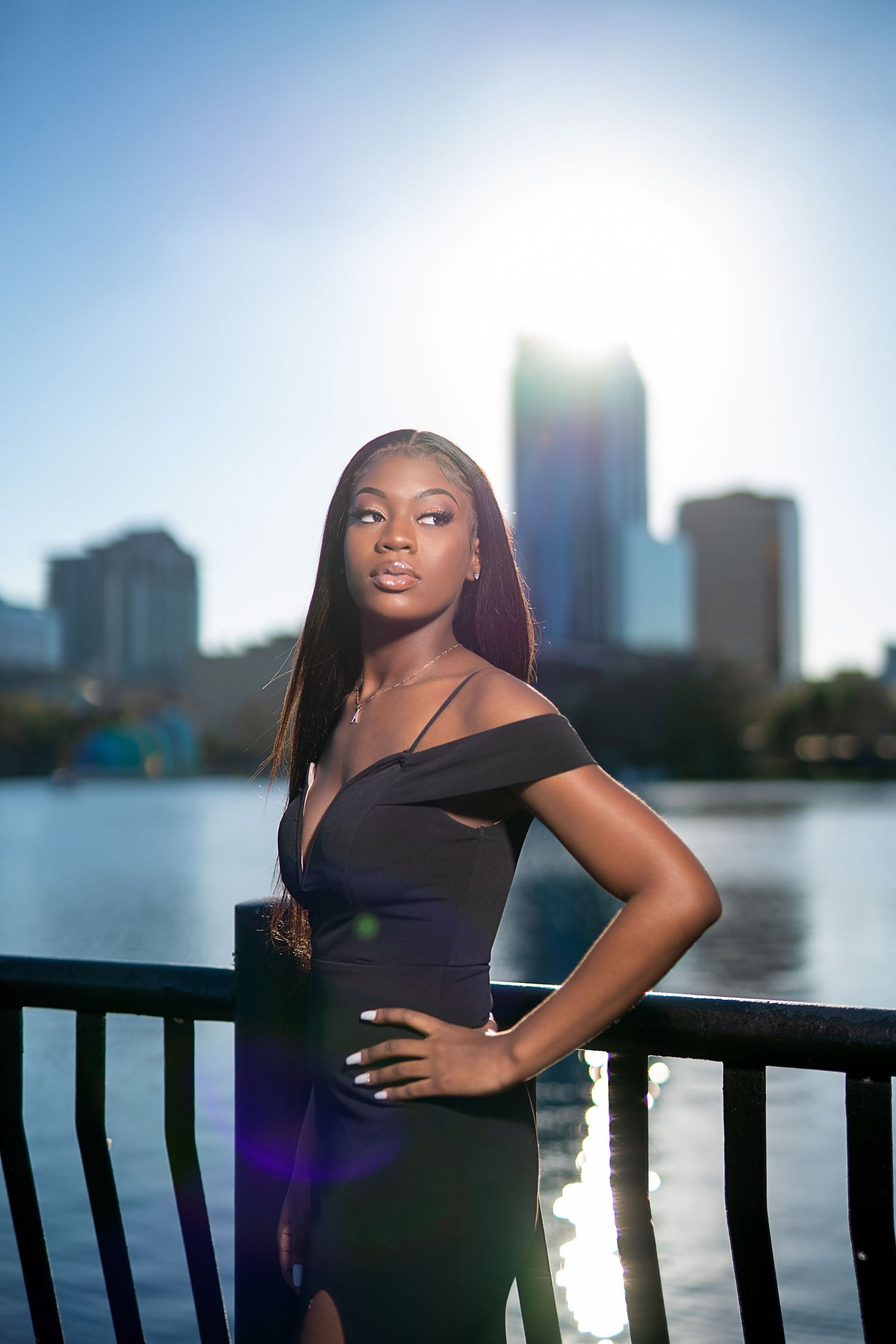 Young Black woman in elegant off-shoulder black dress poses by waterfront railing with city skyline at sunset.
