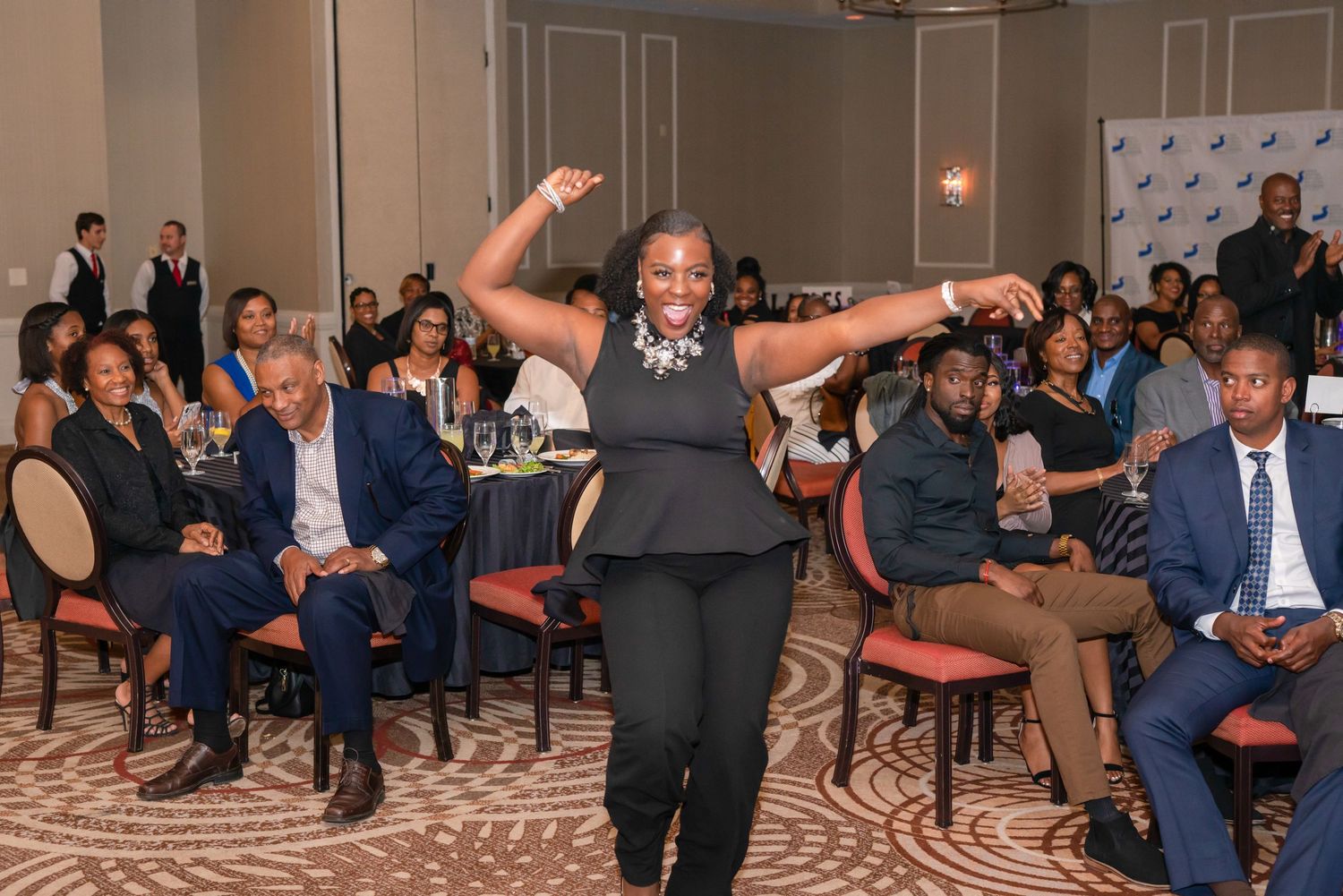 Woman dancing enthusiastically at a formal corporate event with seated audience members watching in a banquet hall.