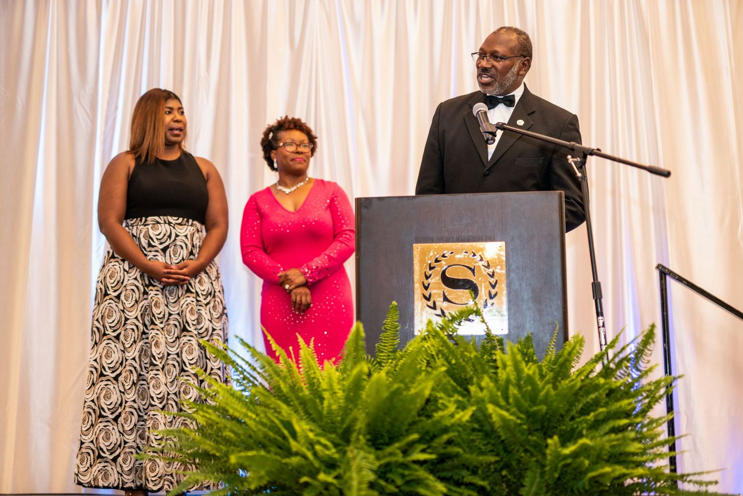 Three people stand at a podium during a formal awards ceremony, with lush green ferns decorating the stage.