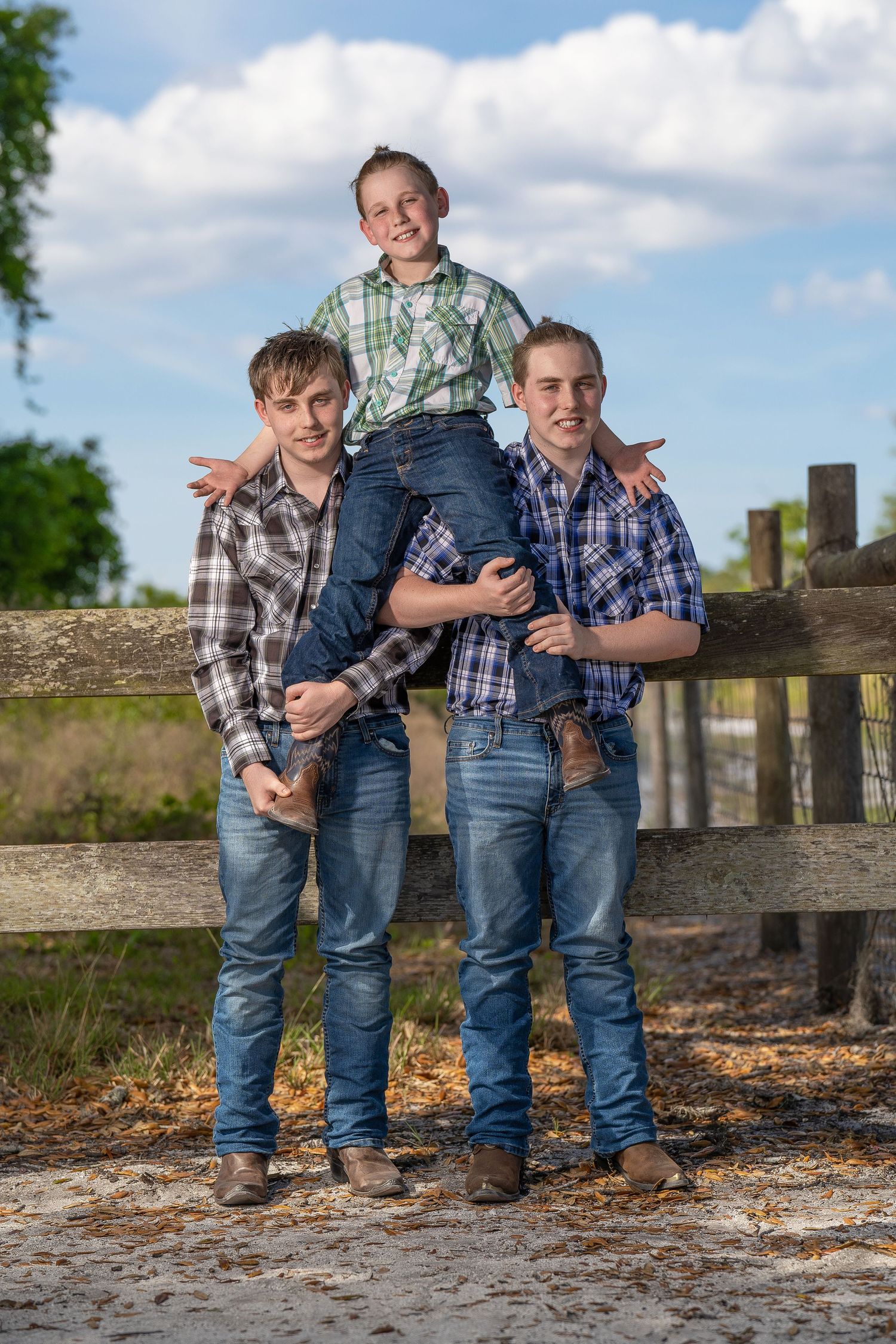 Three boys in plaid shirts and jeans pose by a wooden fence, with the youngest sitting on the older boys shoulders.