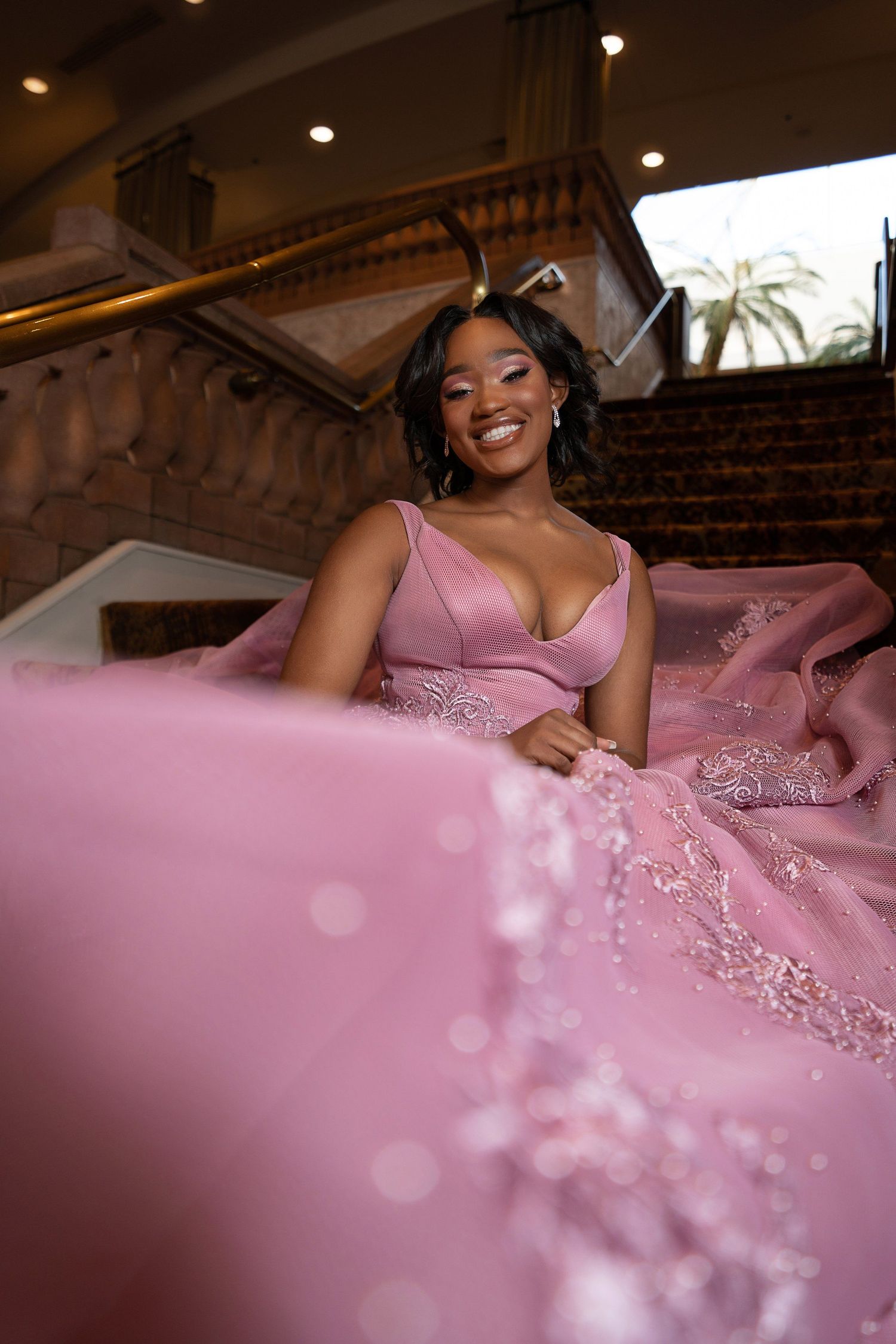 A young woman in a stunning pink ball gown with lace details sits gracefully on elegant stairs, smiling beautifully.