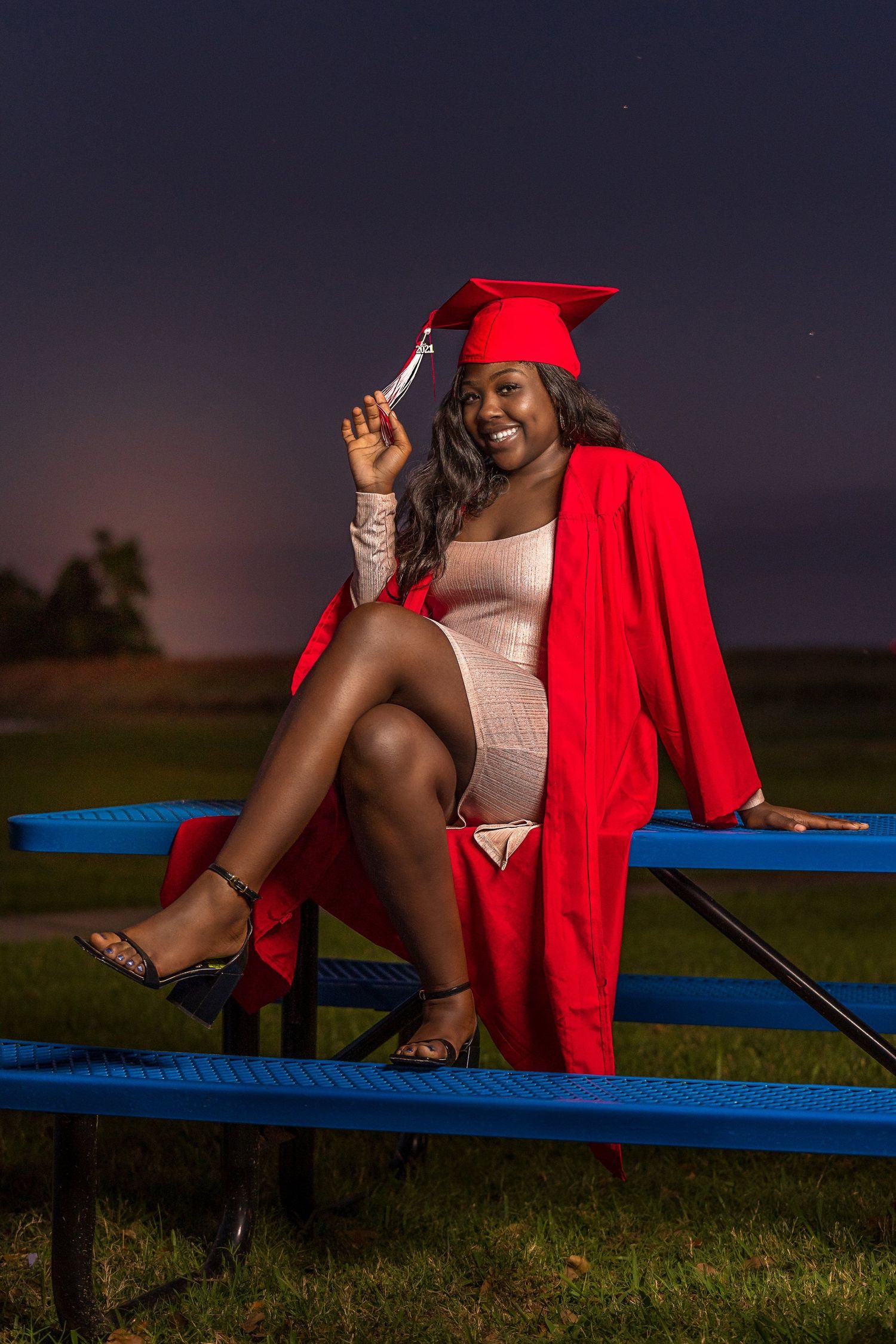 Graduation photos on blue bench with red cap and gown at dusk.