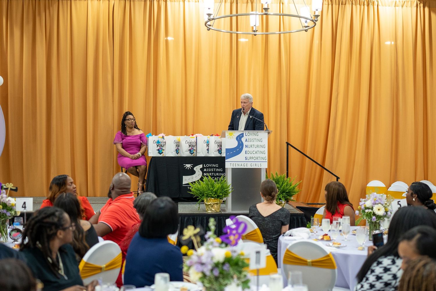 Speaker at podium addresses diverse audience at formal banquet event with yellow drapes and floral centerpieces.