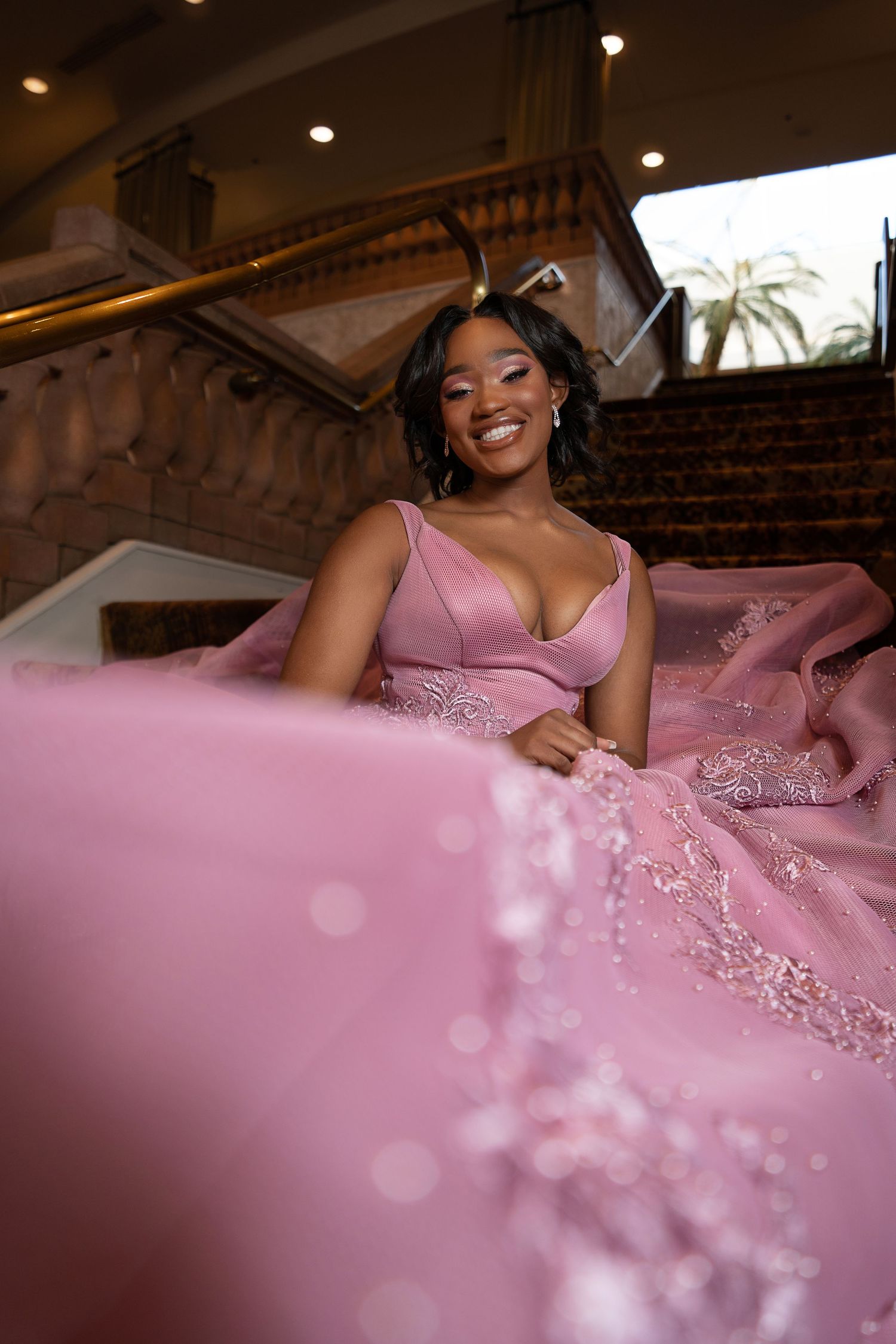 Young woman in elegant pink ballgown with lace details smiles on grand staircase in formal venue.
