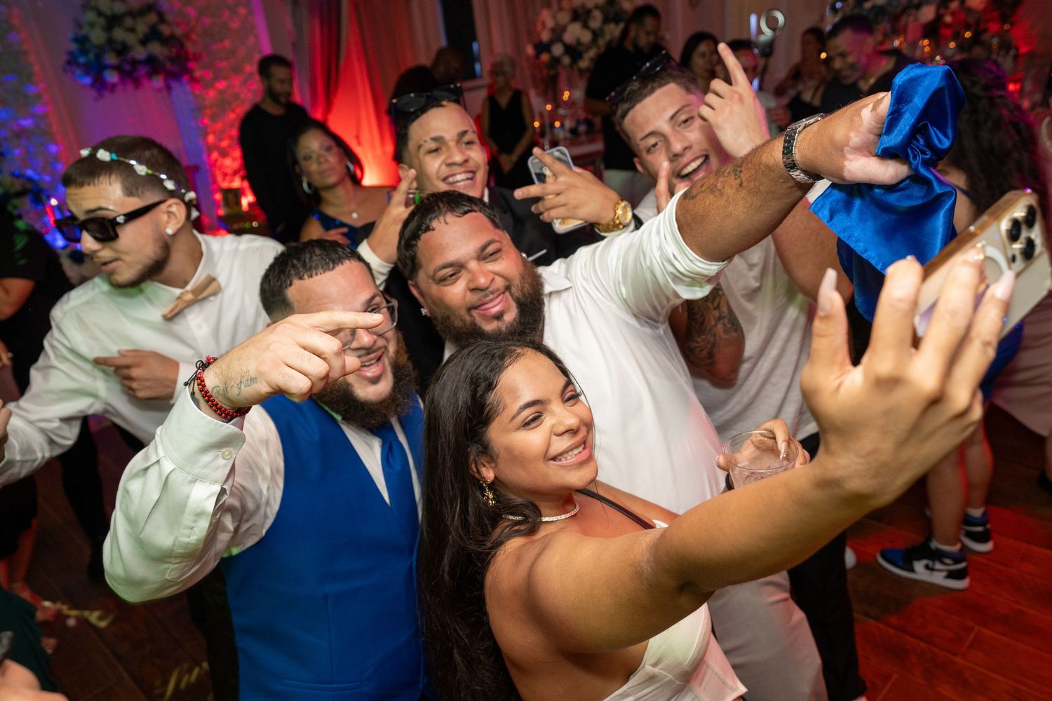 Bride takes a joyful selfie with wedding guests dancing and celebrating at a vibrant reception party.