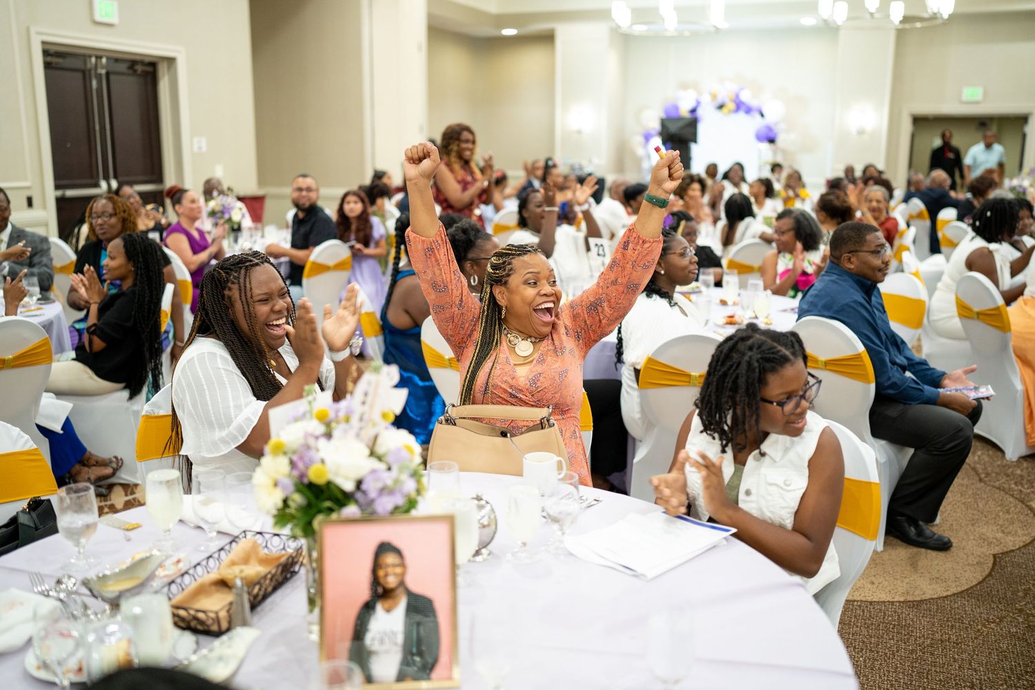 Joyful guests celebrate at an elegant banquet hall event, with a woman raising her arms in excitement.