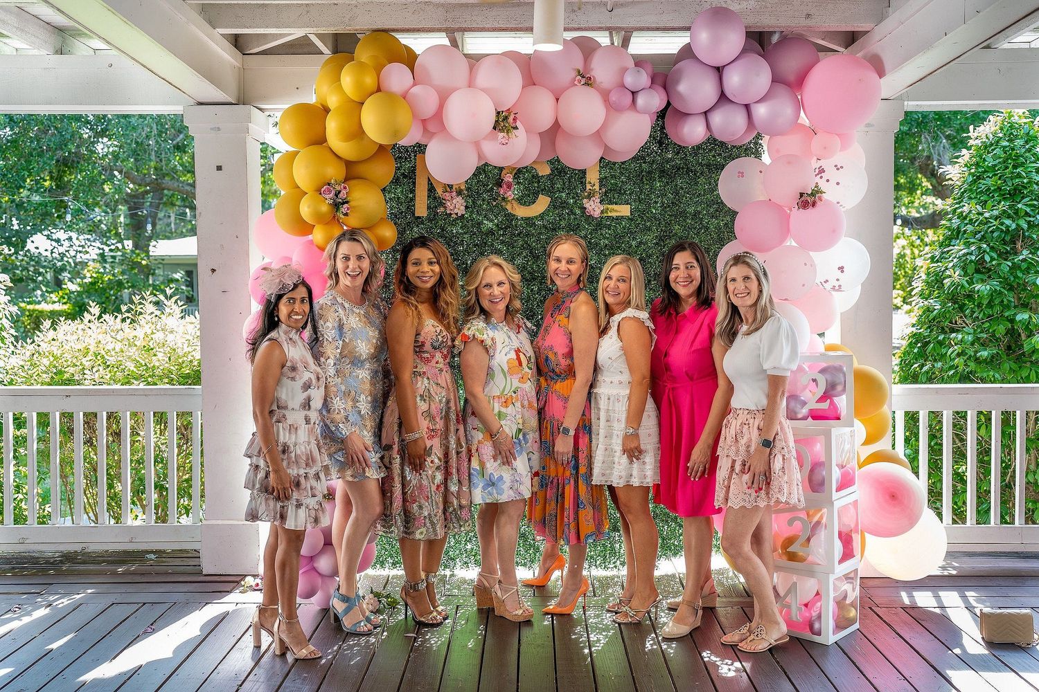 Eight women posing together under a pink and gold balloon arch at an outdoor baby shower celebration.