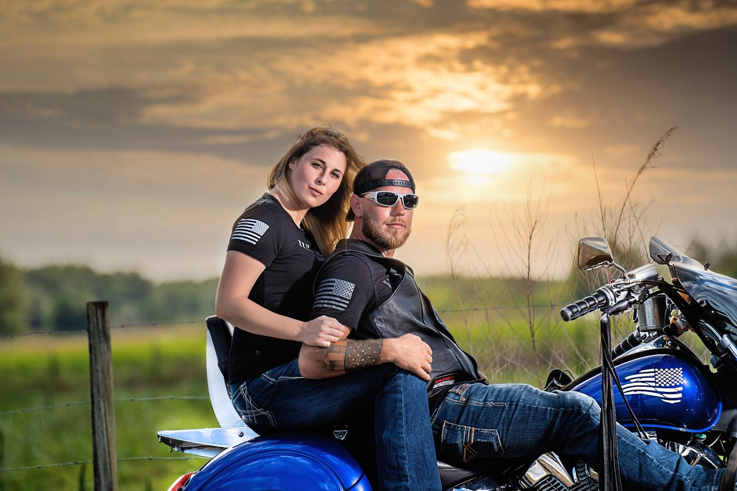 A couple poses on a blue motorcycle at sunset in a rural setting.