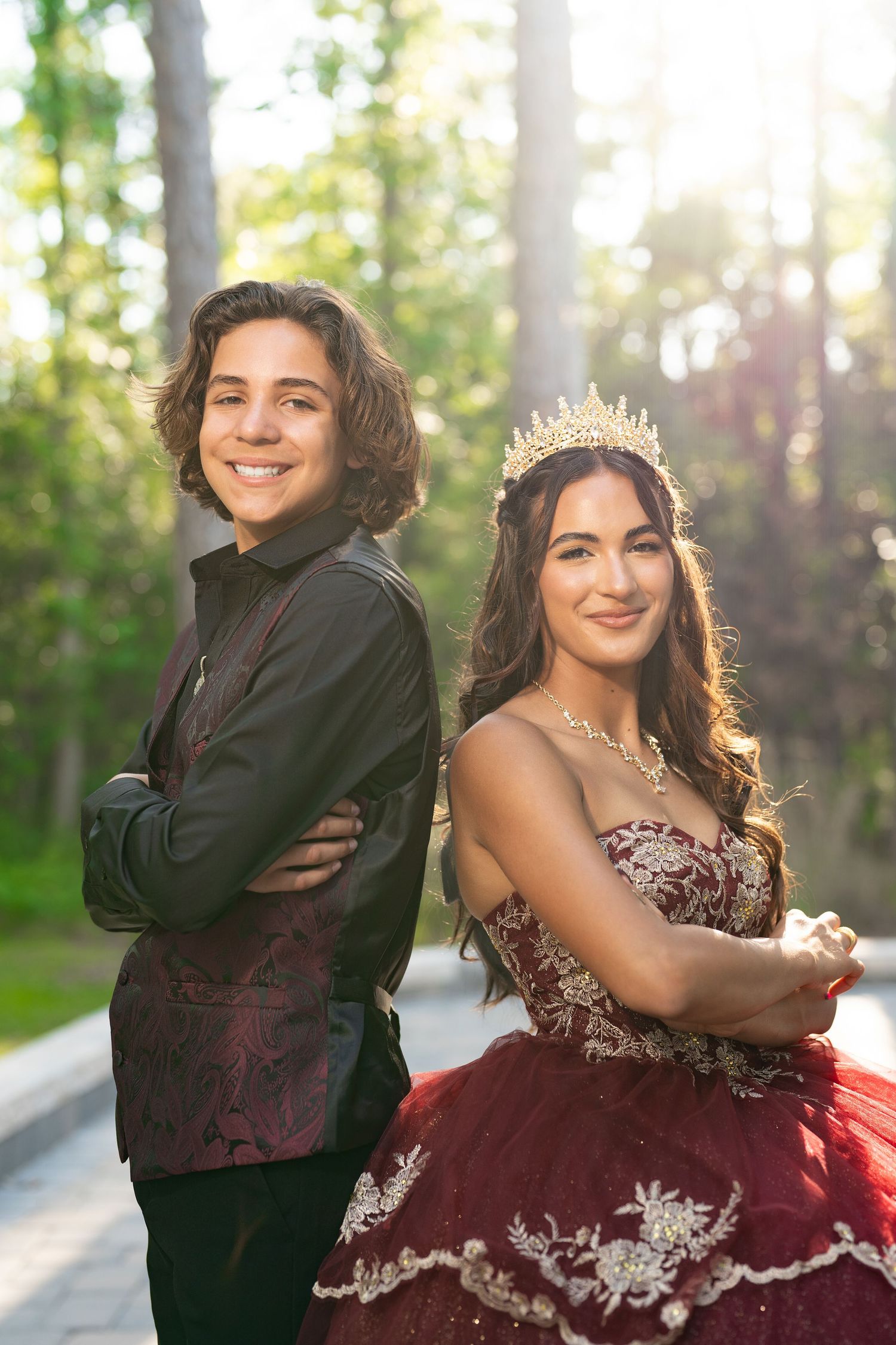 A formal portrait of two people in elegant attire posing in a sunlit forest setting.