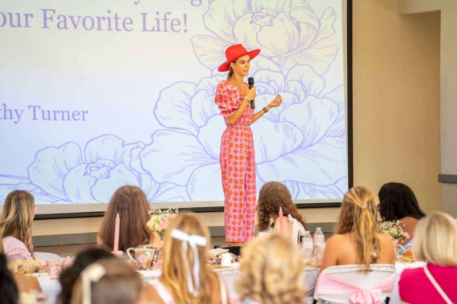 Woman in red floral dress and hat speaks at a live event with a floral presentation slide behind her.