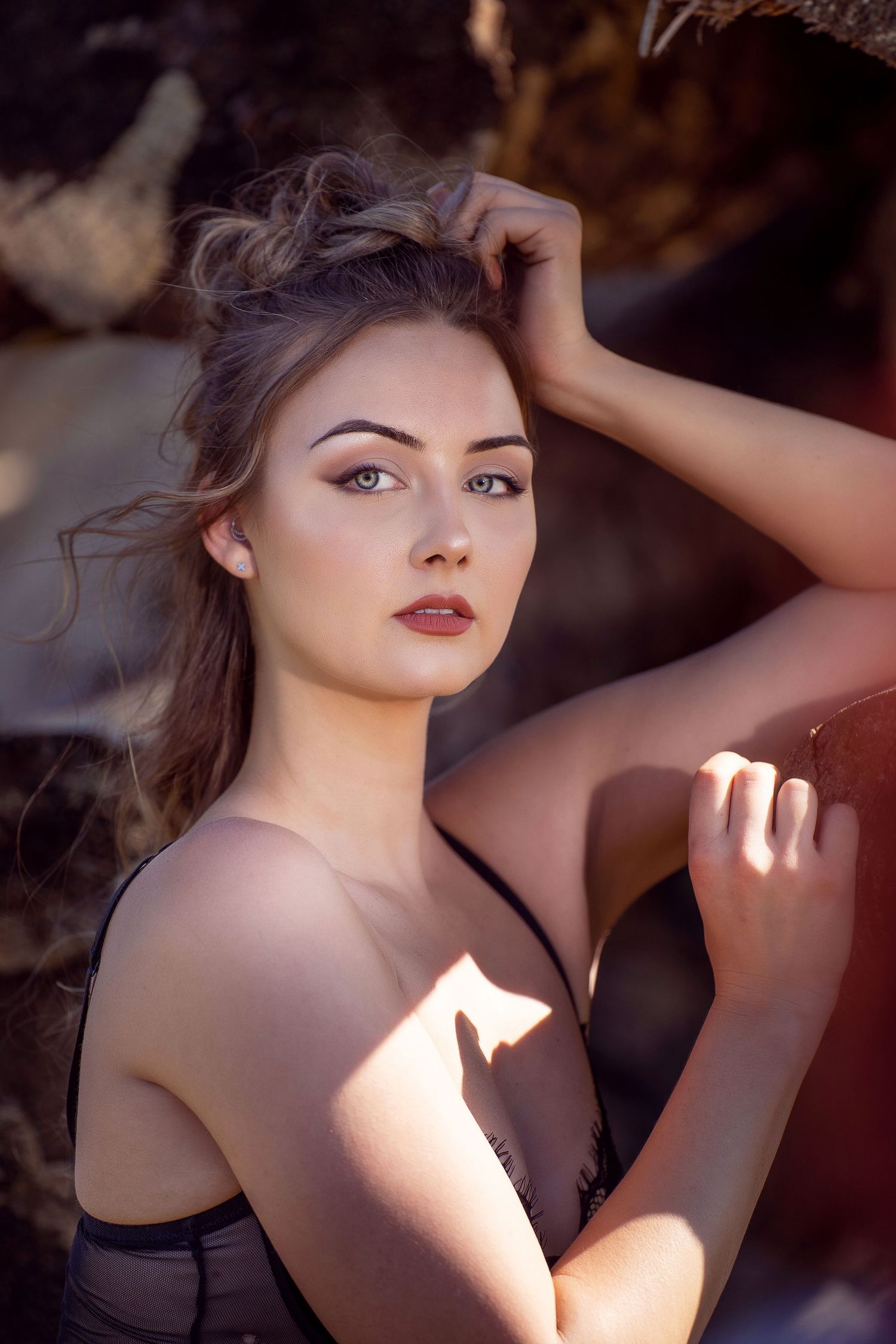 Young woman with updo hairstyle poses gracefully against rocky backdrop in warm sunlight wearing a dark top.