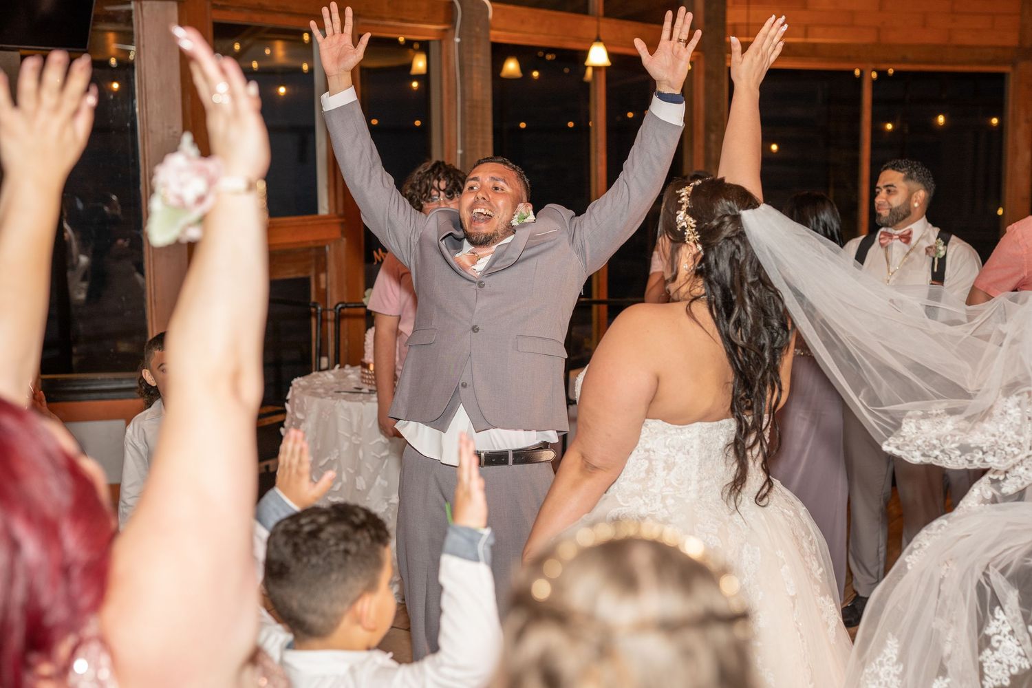 Groom raises hands joyfully at wedding reception as bride and guests celebrate on the dance floor.