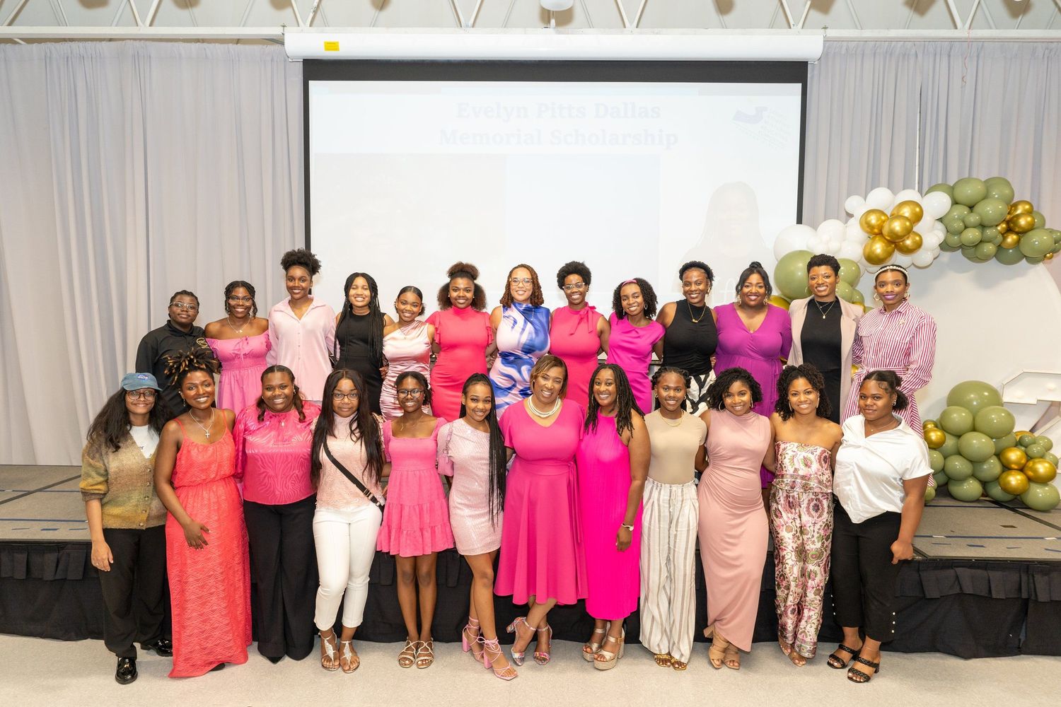 Large group of women in pink and colorful outfits posing together at a formal event with balloon decorations.