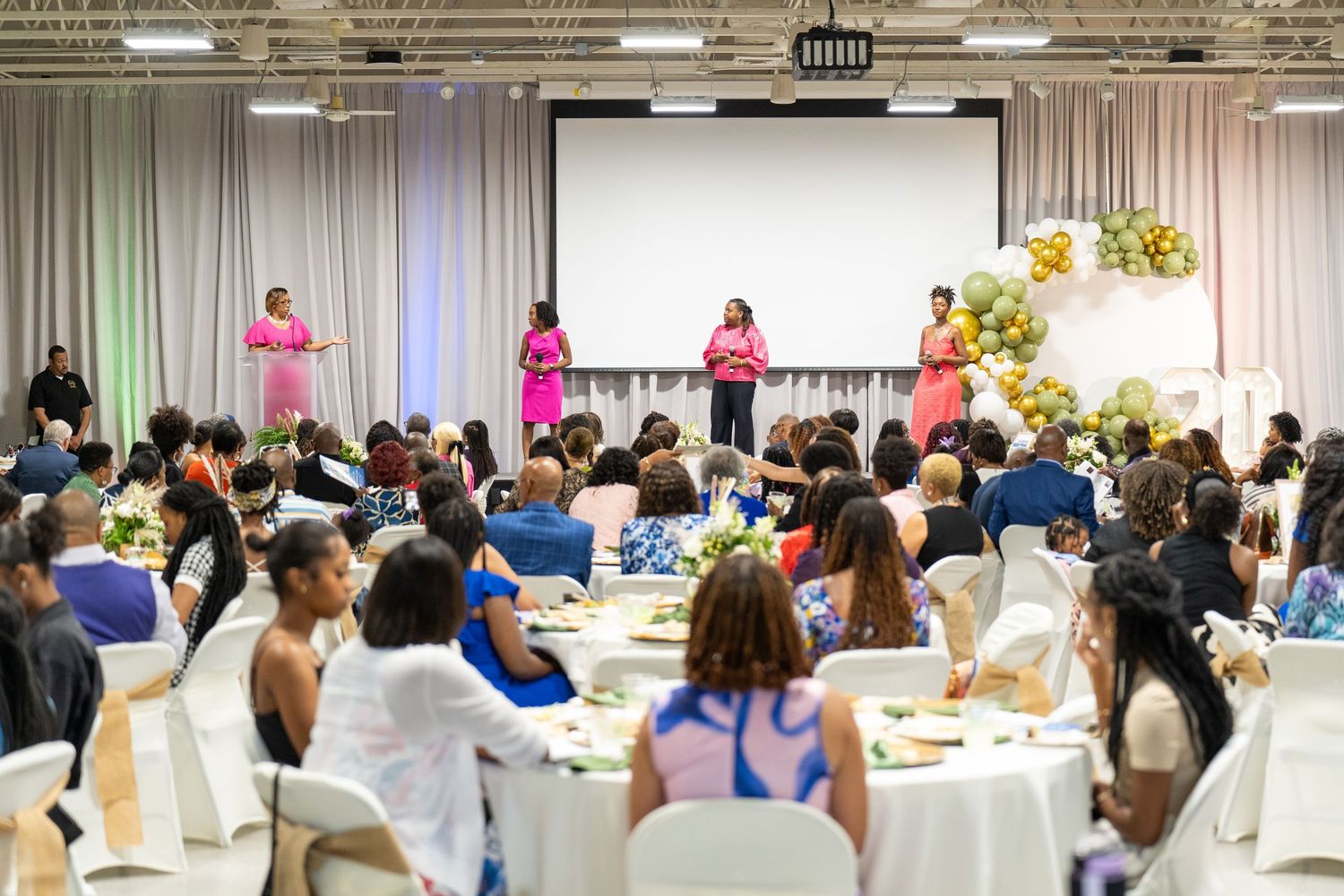 Elegant banquet hall with diverse women seated at round tables during a formal gala event with speakers on stage.