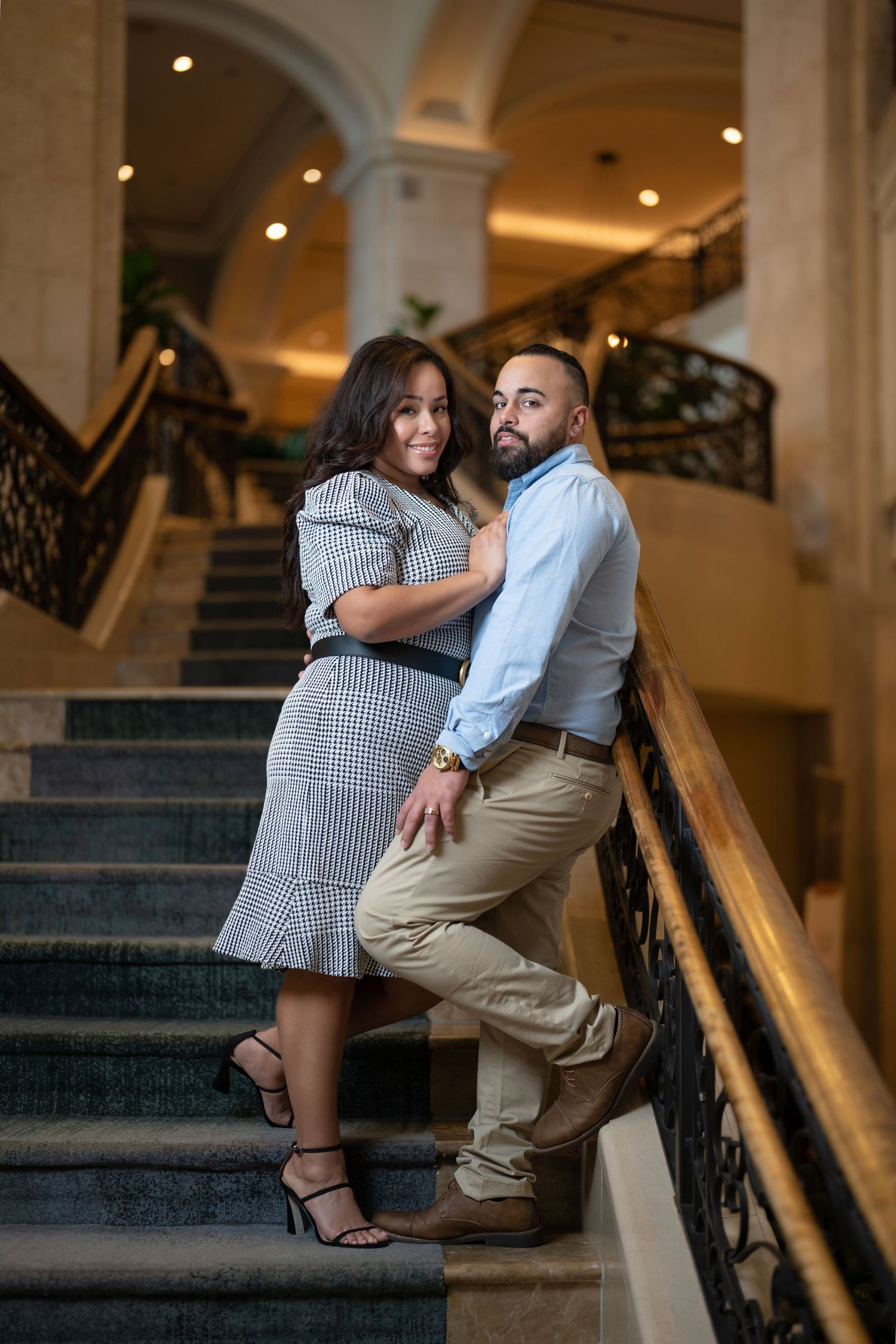 Couple posing elegantly on ornate staircase, woman in plaid dress and man in light blue shirt, in a grand interior.