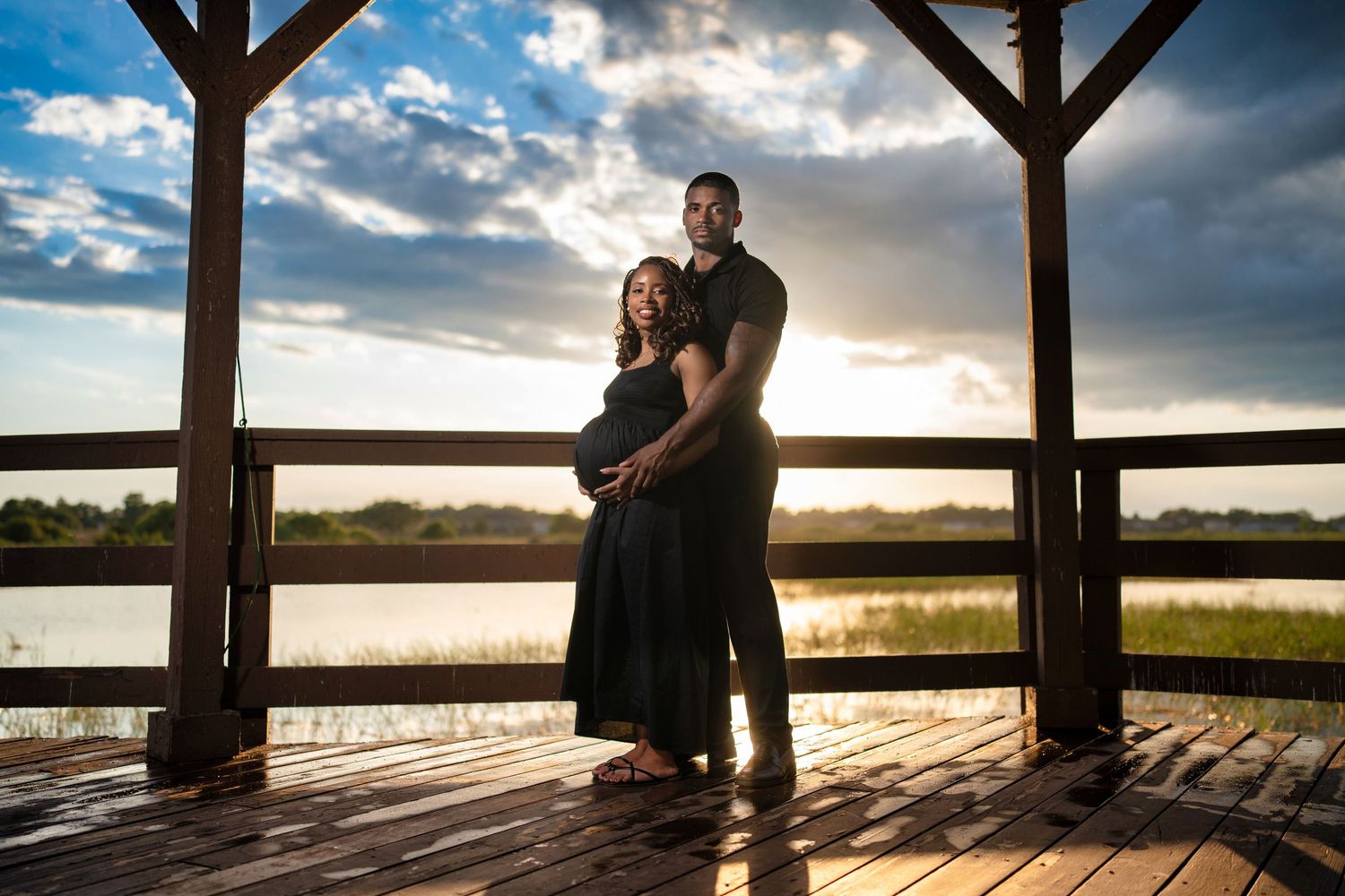 Couple poses on wooden deck at sunset, woman in black maternity dress, man embracing her from behind, lake in background.