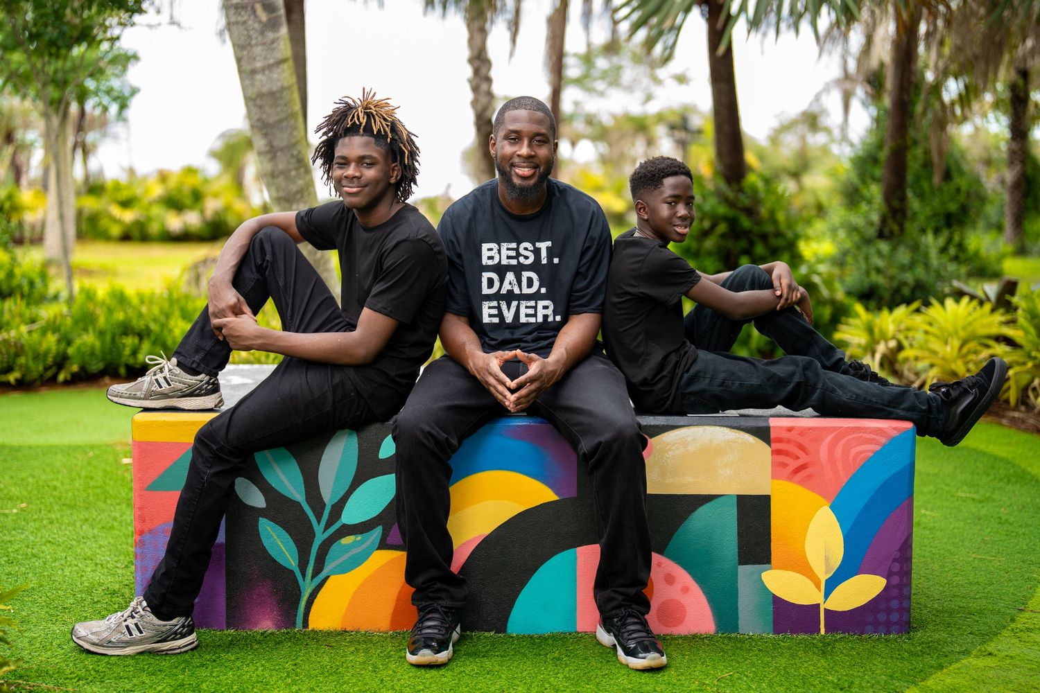 A father wearing a Best Dad Ever shirt poses proudly with his two sons on a colorful bench outdoors.