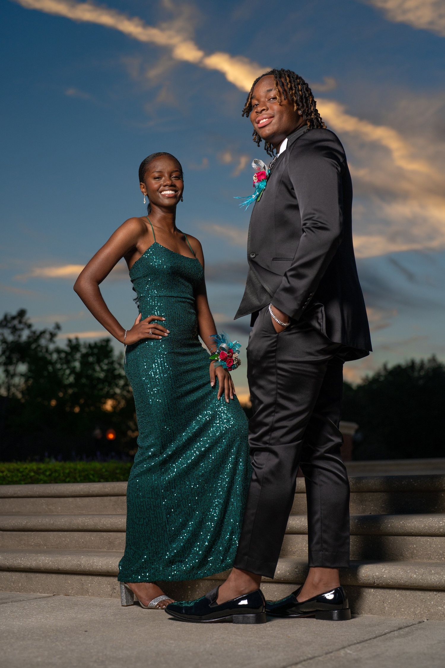 A young couple dressed in prom attire, woman in green sequin gown and man in black suit, posing outdoors at sunset.