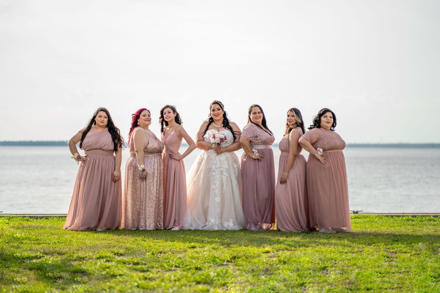 Bride in white lace gown poses with six bridesmaids in dusty rose dresses by a waterfront on a sunny day.