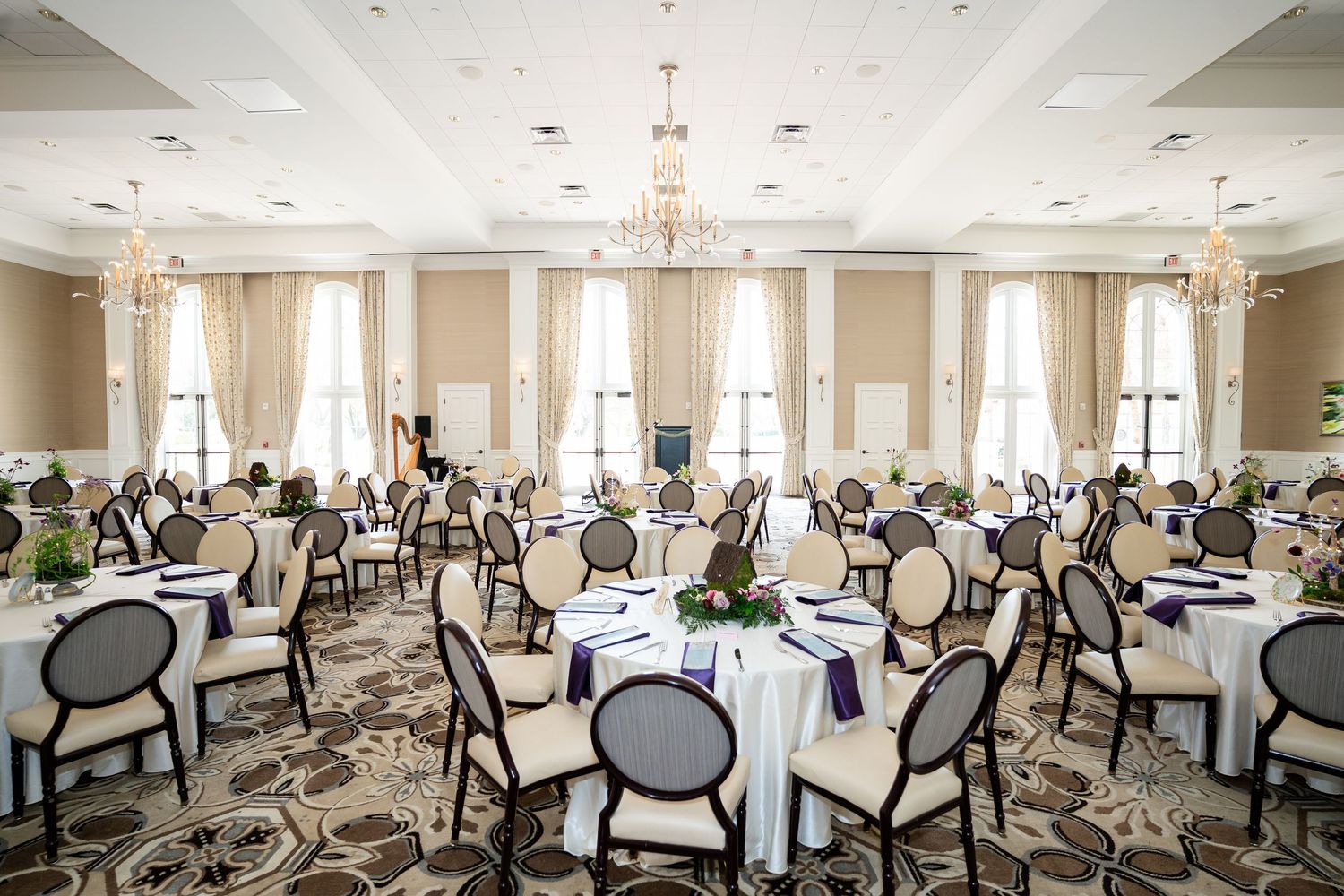 Elegant ballroom with round tables set for a wedding reception, featuring white linens, purple accents, and ornate chandelier lighting.