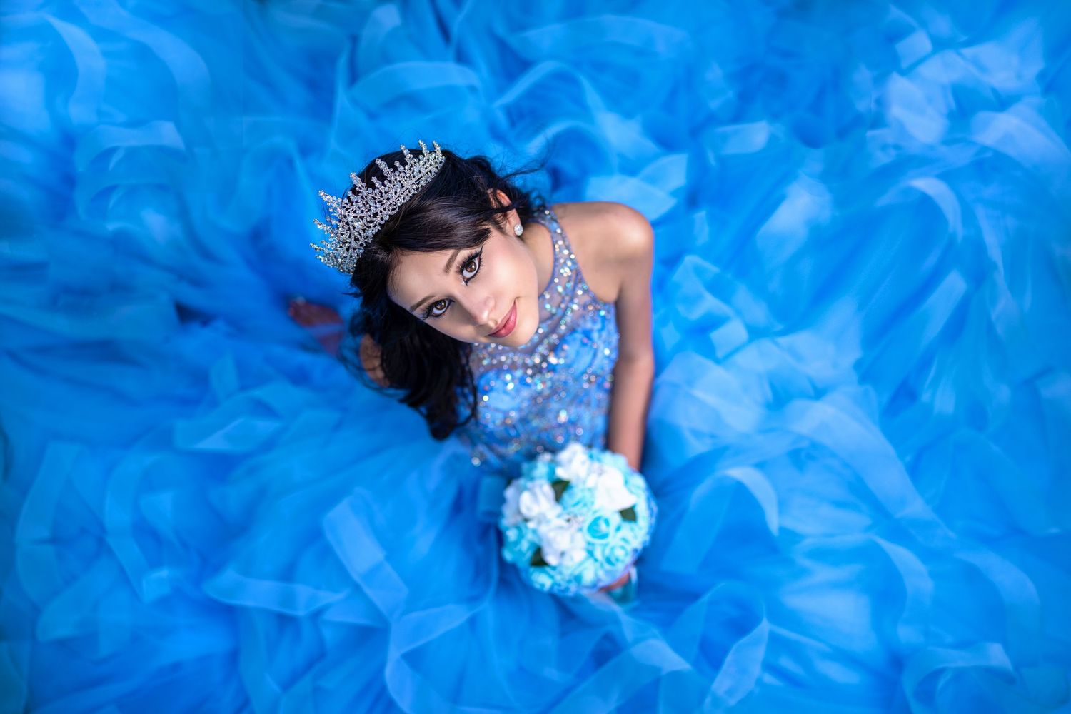 Young girl in a stunning blue ball gown with tiara holds a bouquet, looking up in a dreamy quinceañera portrait.
