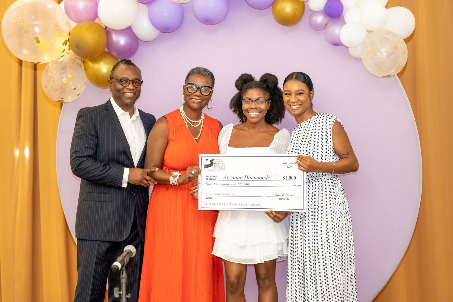 Four people pose with a large scholarship check at a celebration event with balloon arch backdrop.