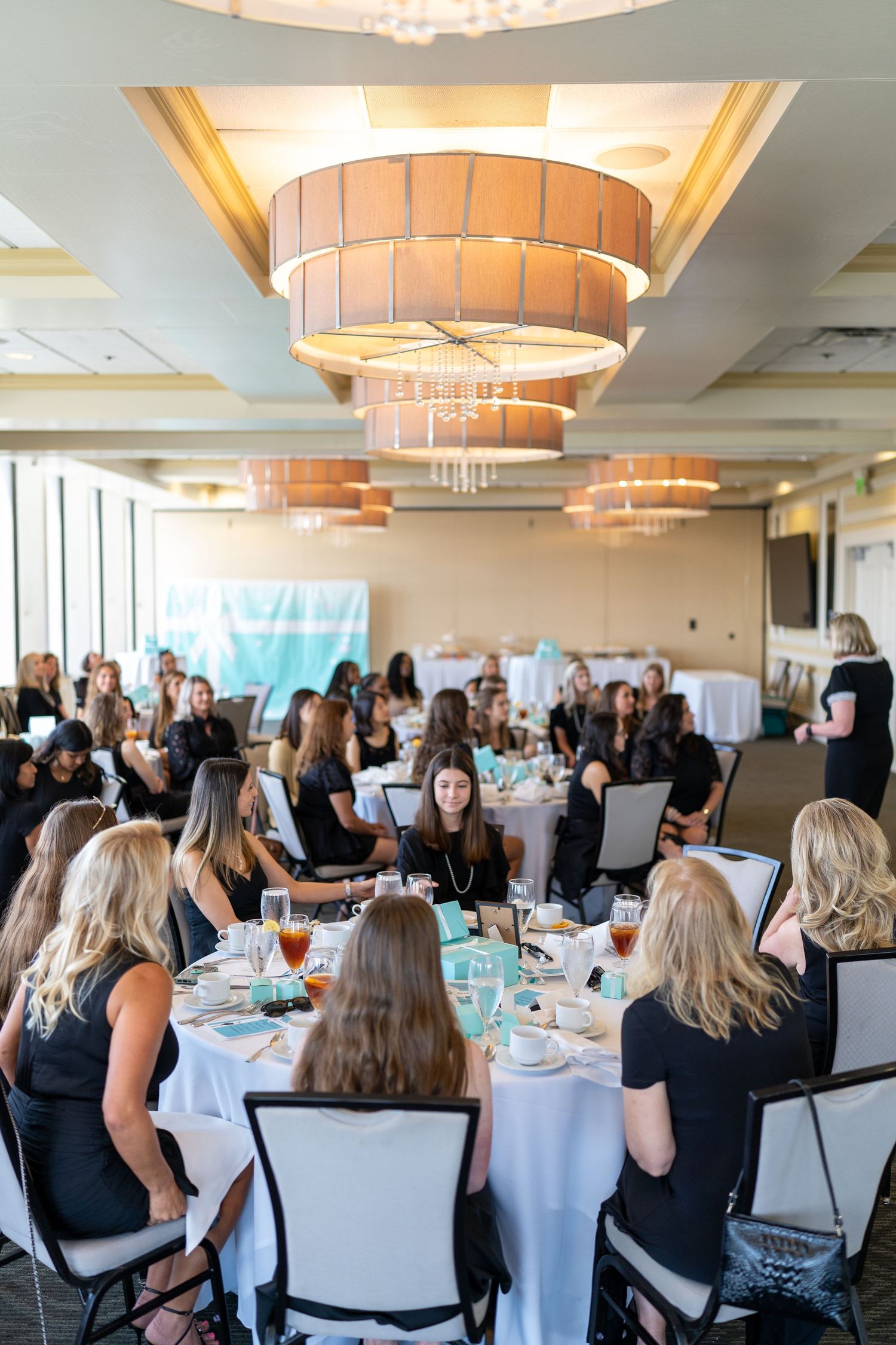 Women in black attire seated at round tables during an elegant banquet event in a ballroom with decorative chandeliers.