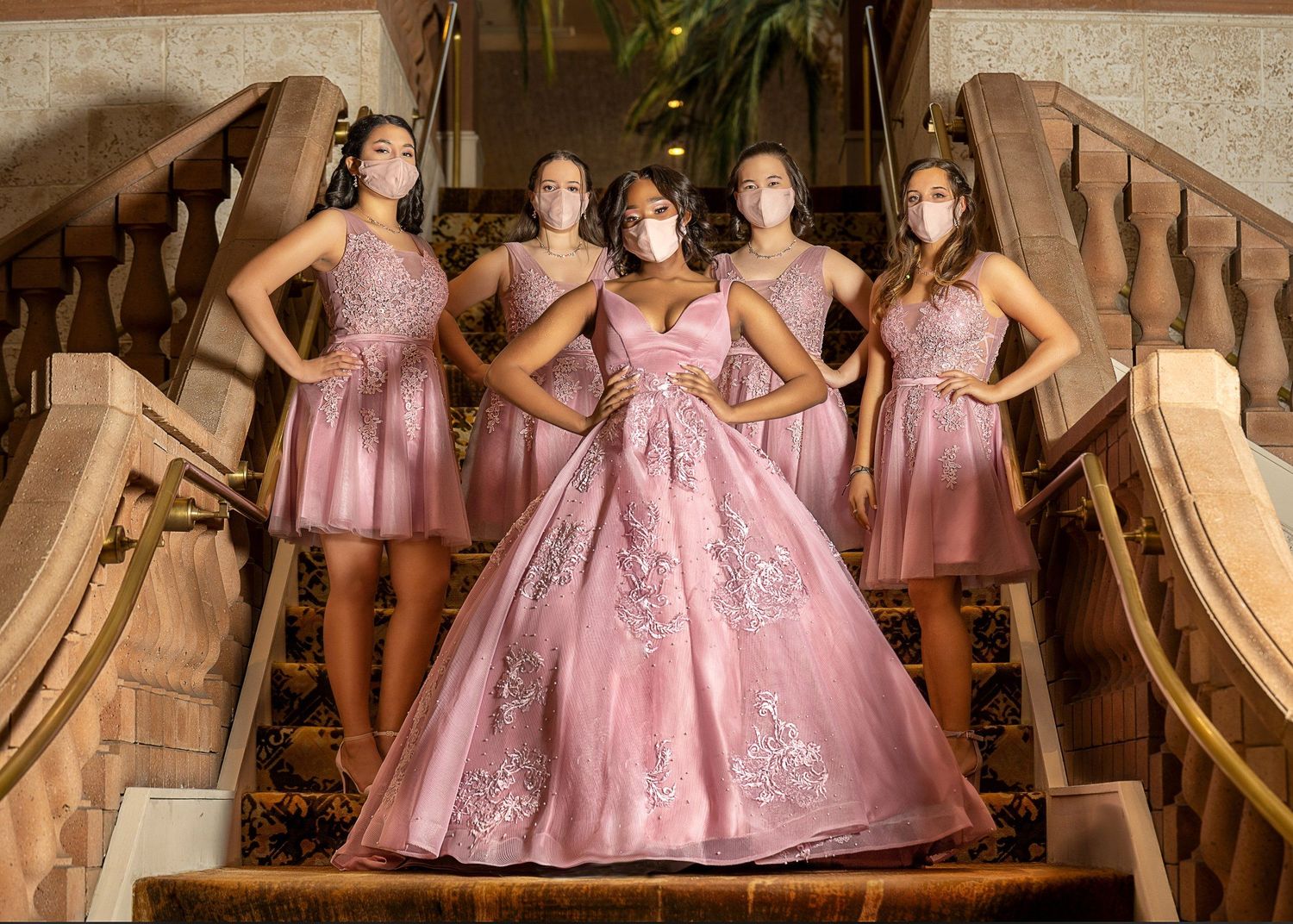 Five young women in pink dresses and masks pose on ornate staircase during a quinceañera celebration.