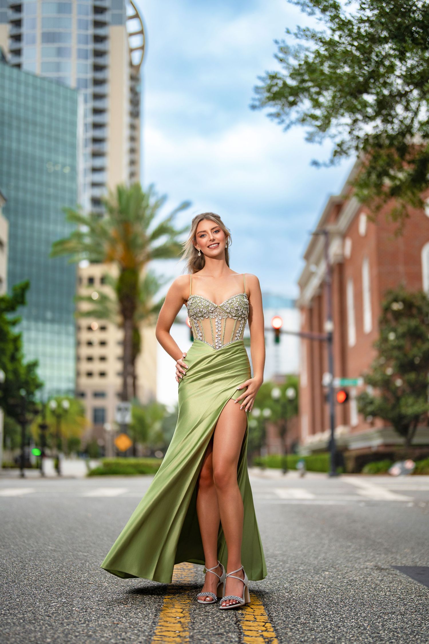 Woman in green satin gown with embellished bodice and thigh slit poses on urban street with city buildings backdrop.