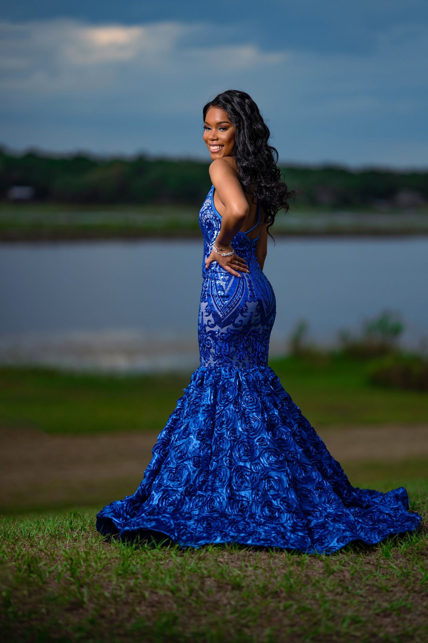 Woman in blue sequined mermaid gown poses by lake during sunset.