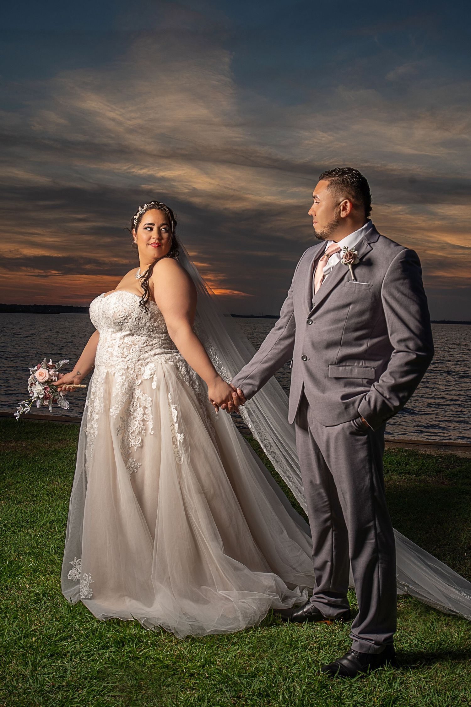 Wedding couple poses on grass by waterfront during dramatic sunset.