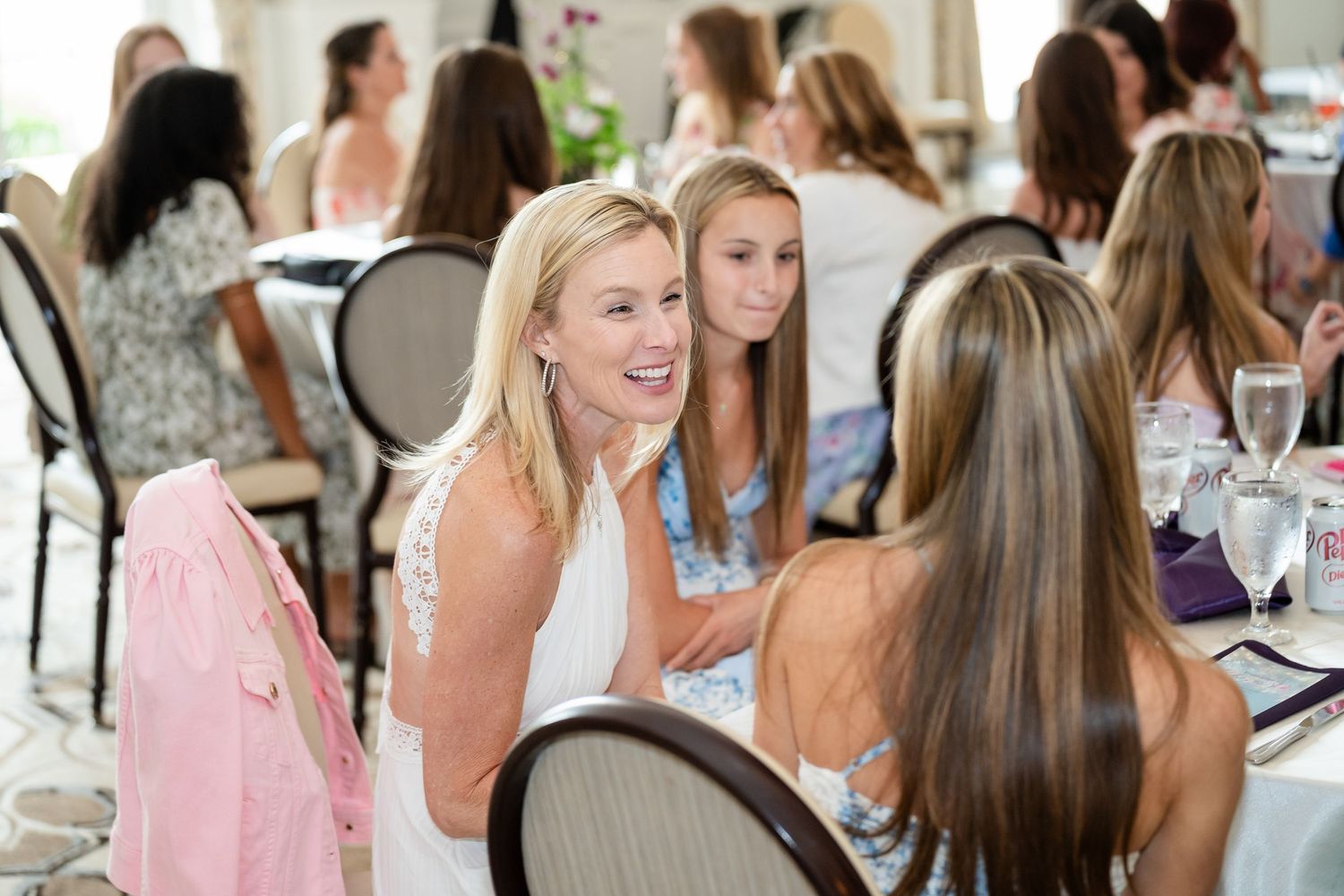 Women socializing at an elegant indoor event, smiling and conversing at decorated tables in a bright venue.