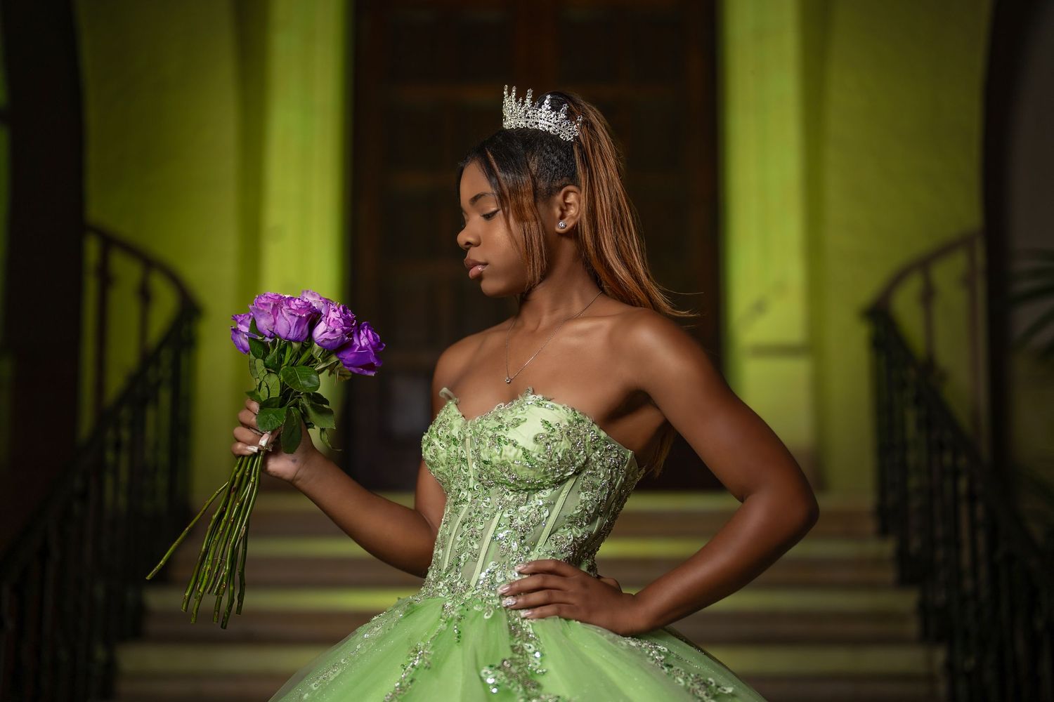 Young Black woman in green ball gown with tiara holds purple flowers on ornate staircase.