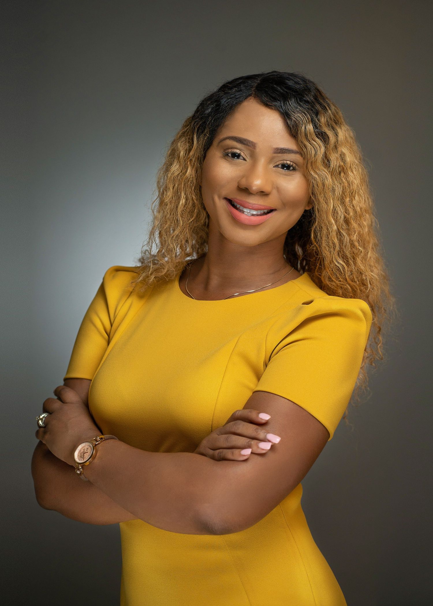 Professional woman in yellow dress with crossed arms, smiling confidently against gray background.