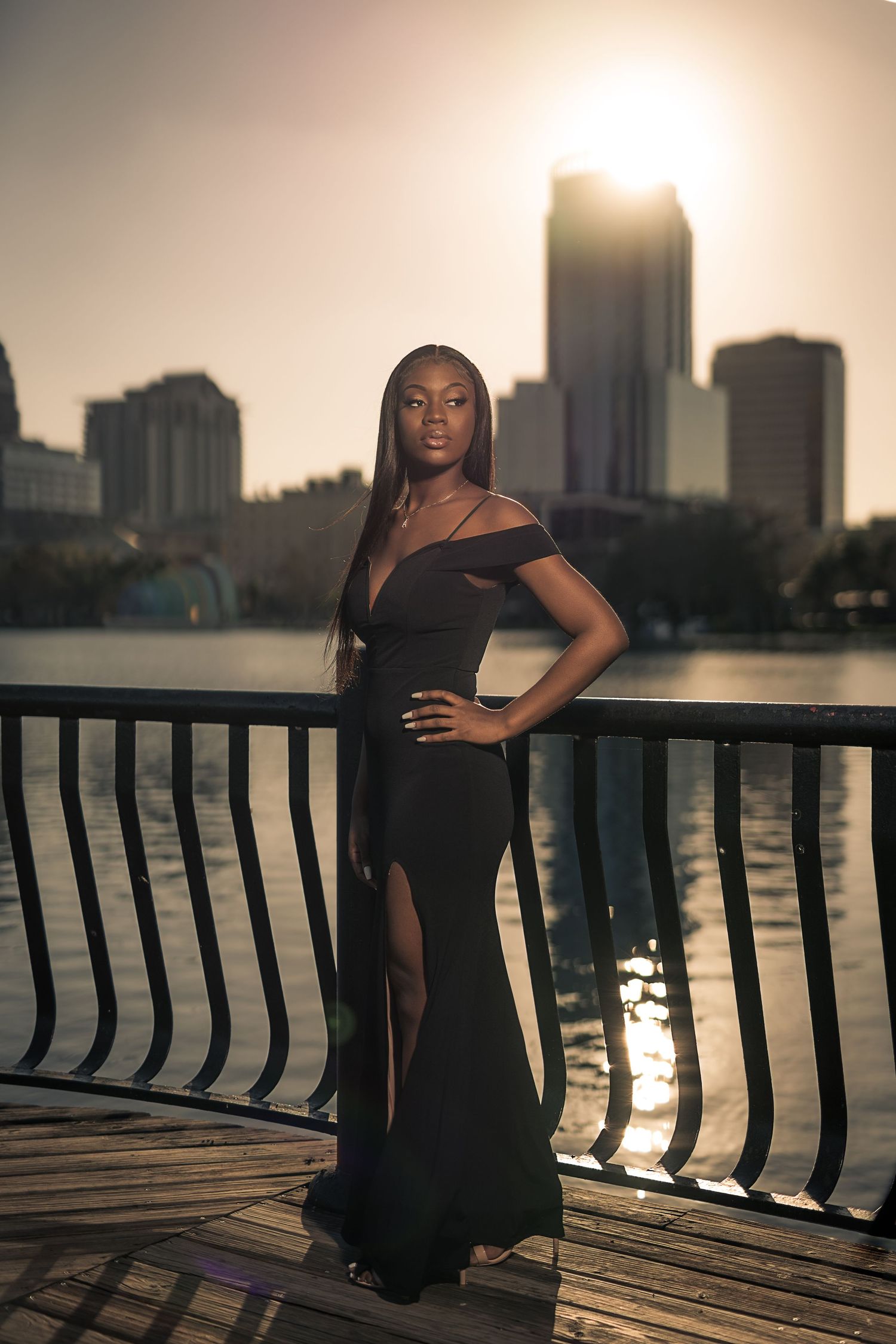 Young woman in elegant black gown poses on waterfront boardwalk at golden sunset with city skyline backdrop.