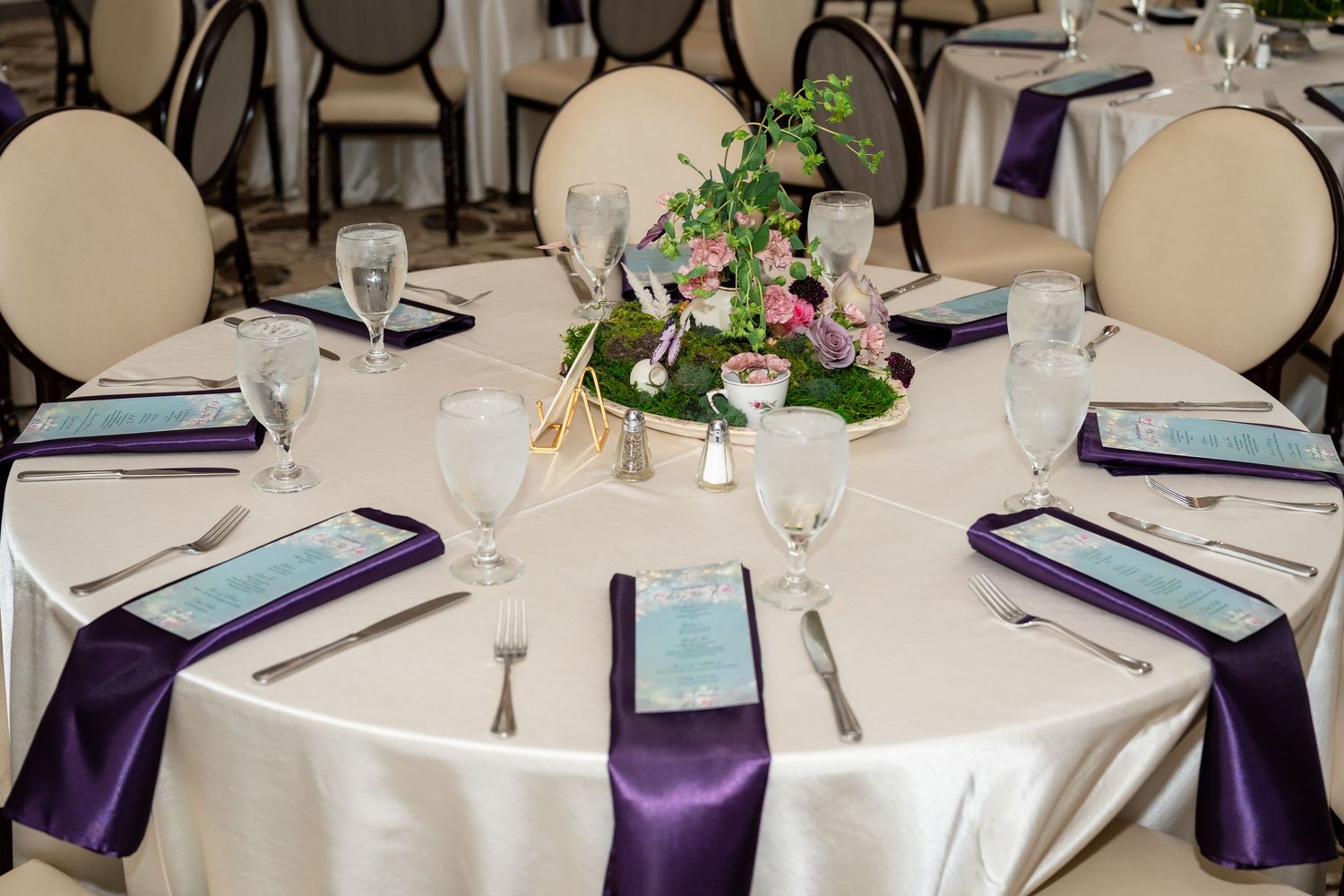 Elegant round banquet table with purple satin napkins, crystal glassware, silver cutlery, and floral centerpiece.