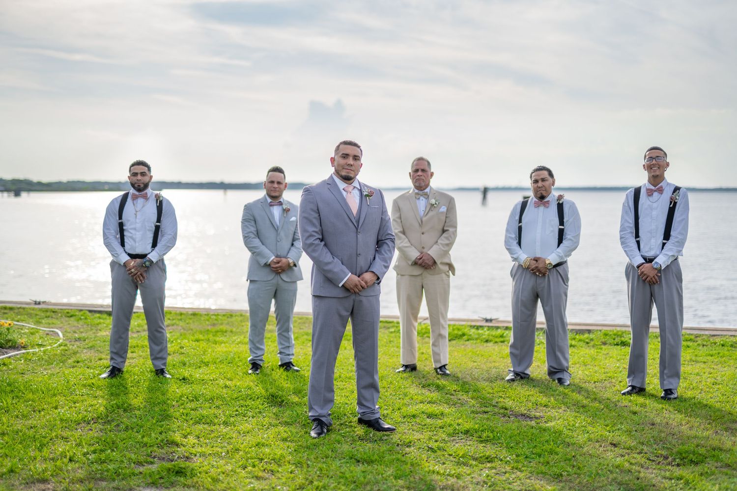 Groom stands in front of groomsmen in gray suits with black suspenders on a waterfront lawn at an outdoor wedding.