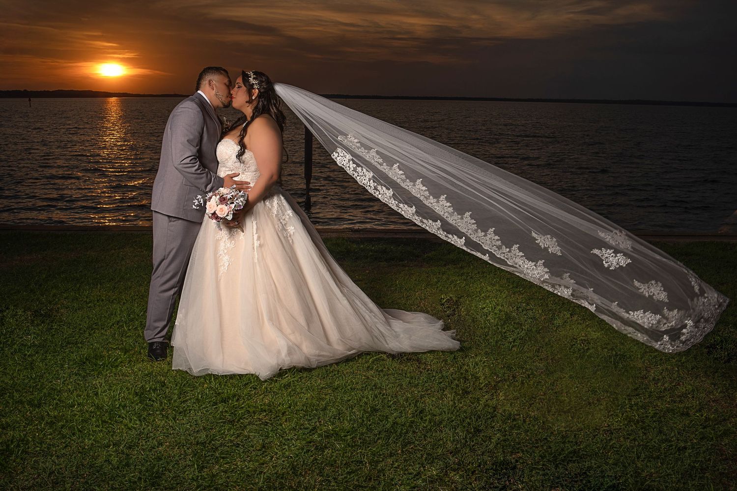 Bride and groom kissing at sunset by the water, with flowing lace veil billowing in the wind.