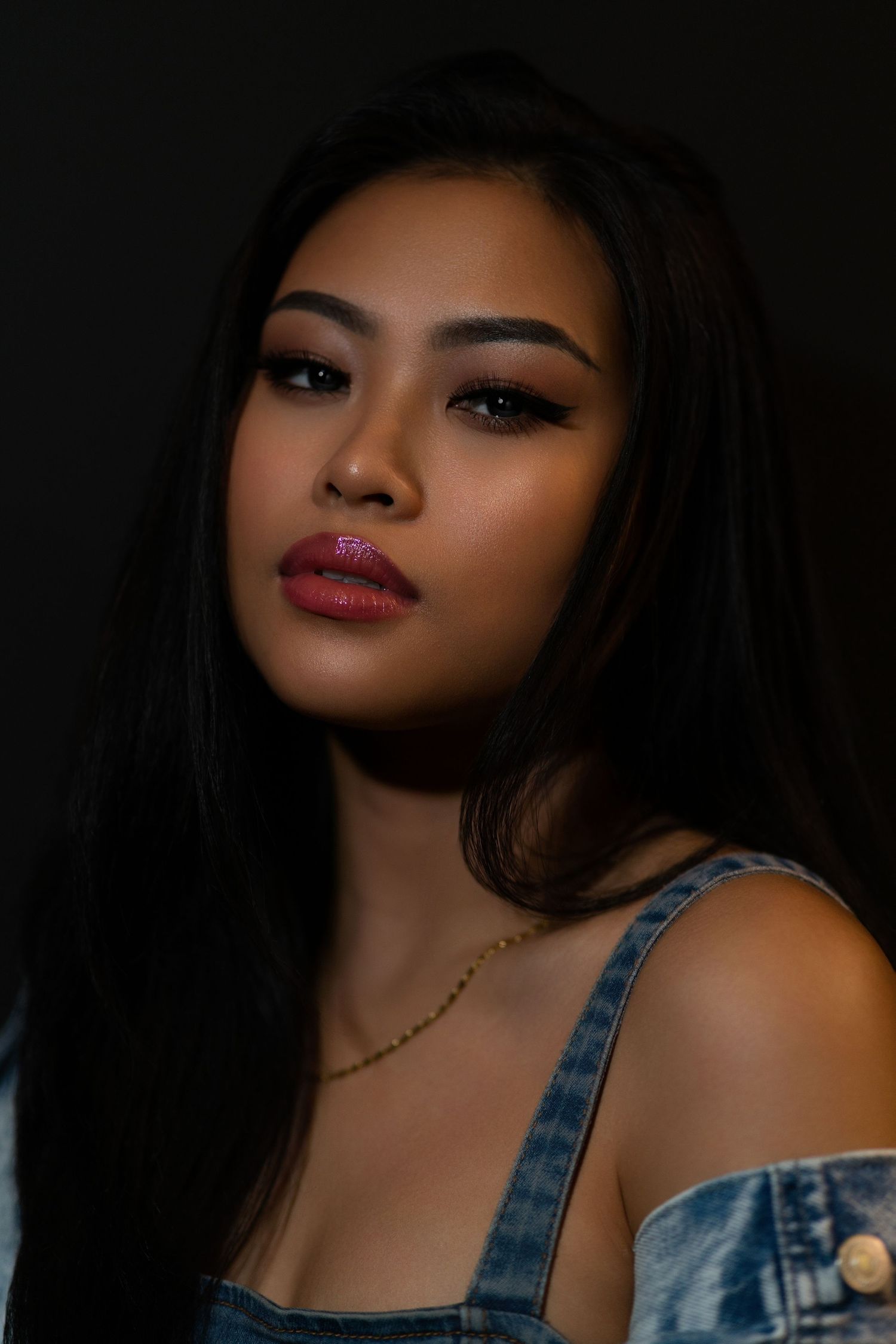 Young Asian woman with long dark hair, glittery pink lips, and gold necklace wearing denim overalls against dark background.