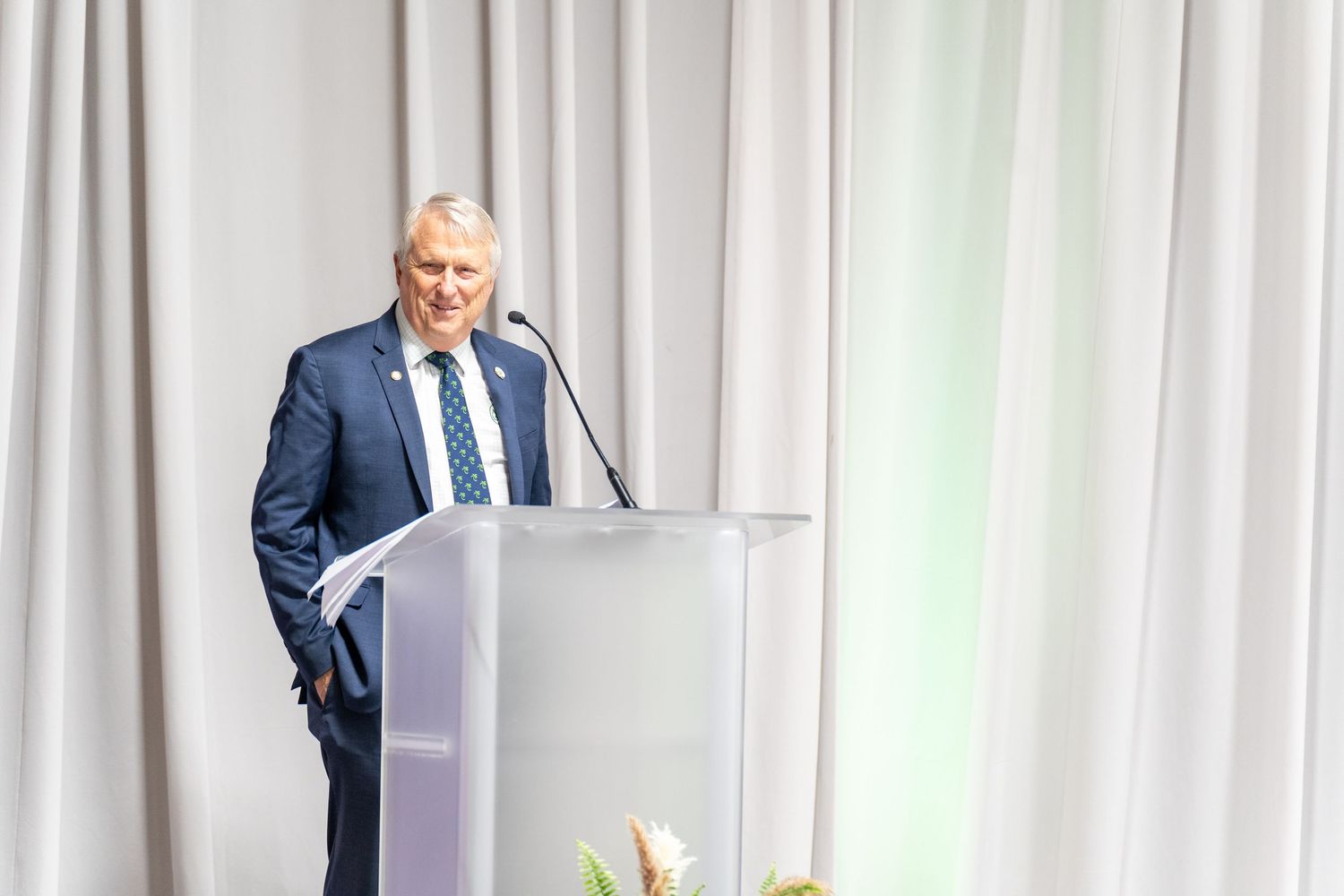 Man in blue suit speaking at a podium with microphone at a formal conference event.