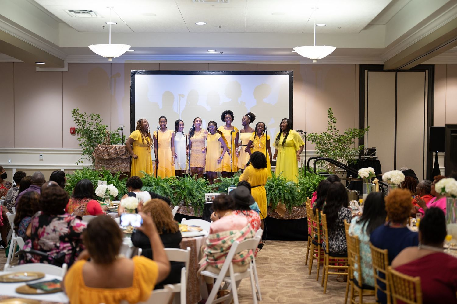 Women in yellow dresses perform on stage at an elegant banquet hall event with seated audience.