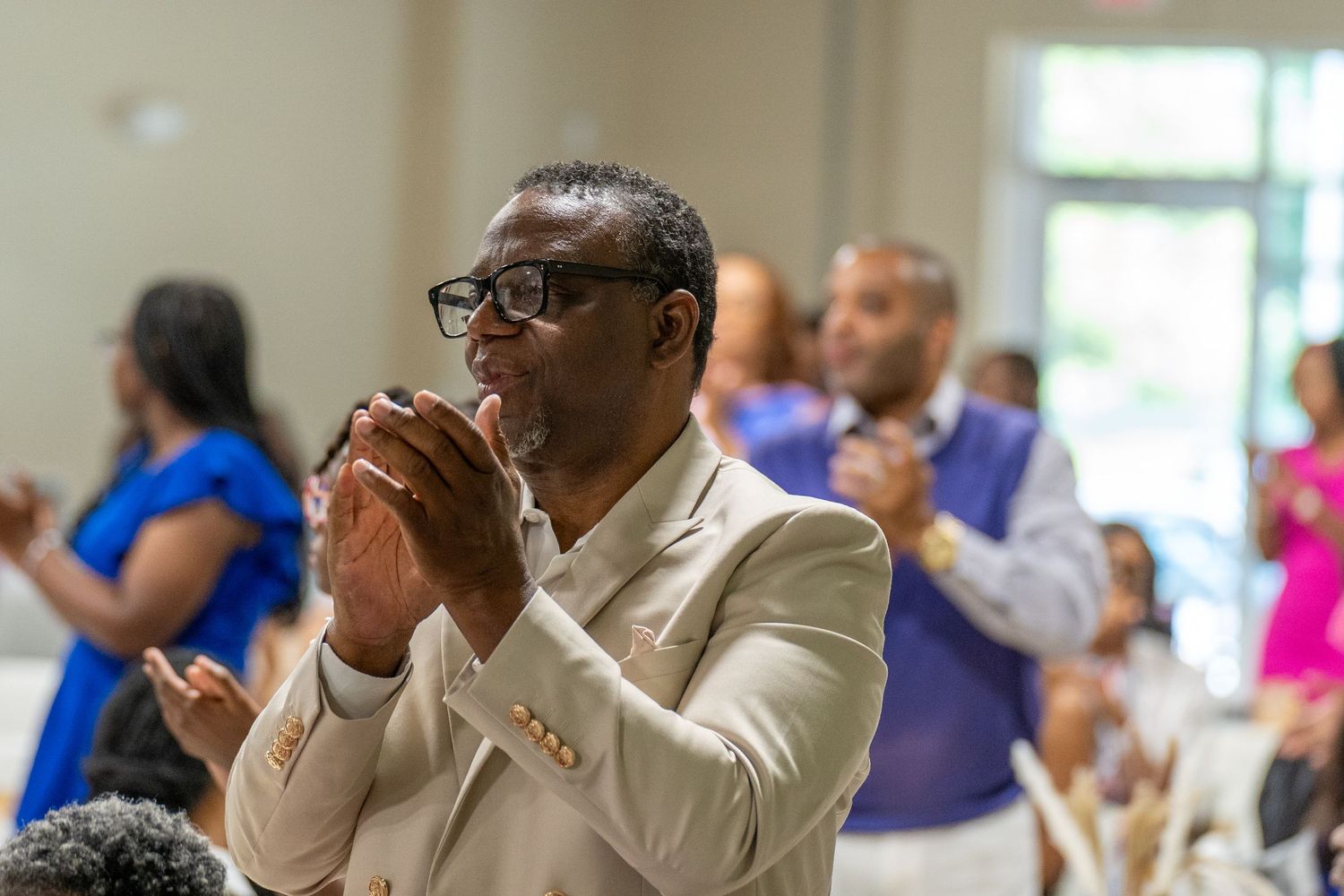 A man in a beige suit claps his hands during a church service with attendees in the background.