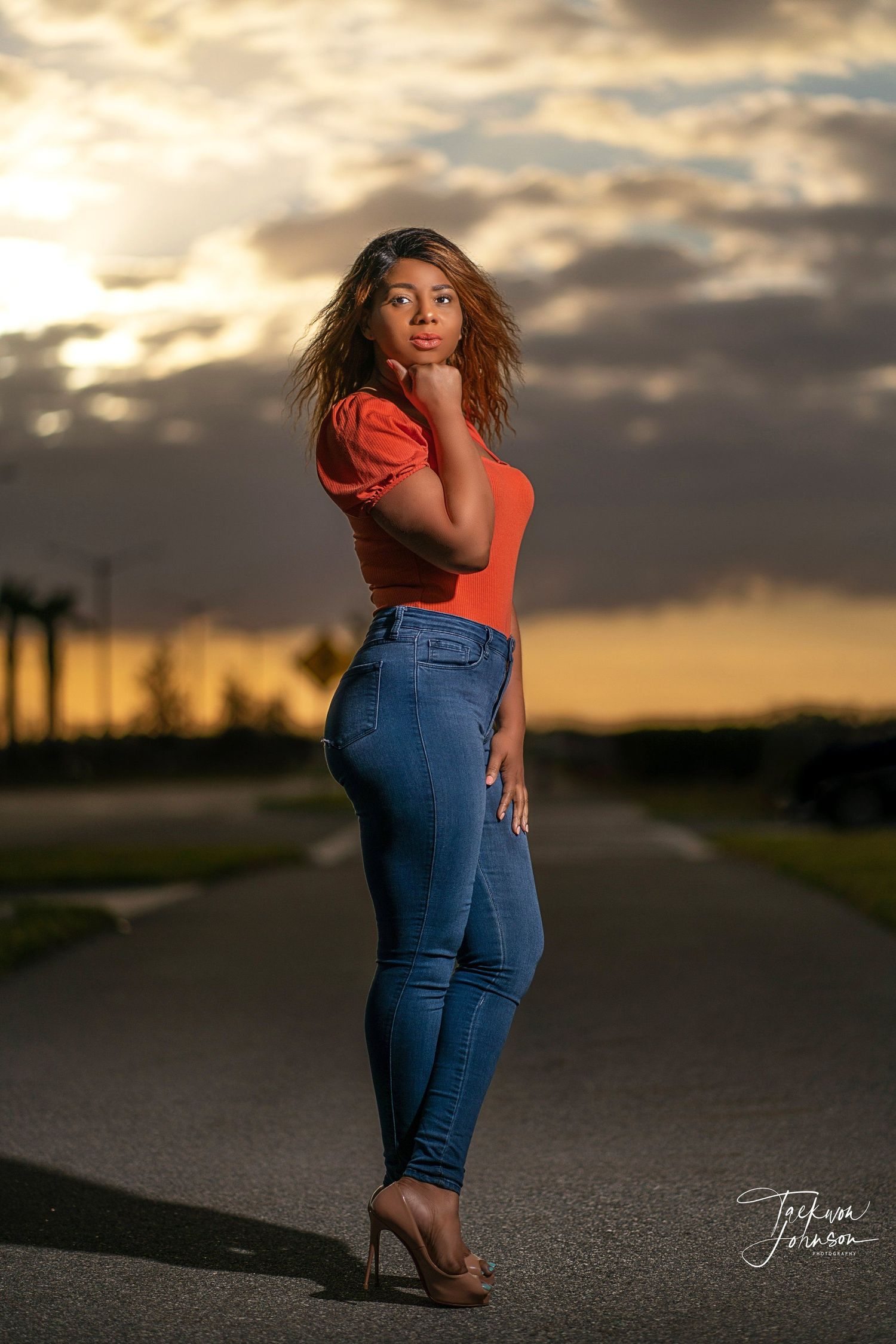 Sunset portrait of a woman in orange top and jeans posing on a road with dramatic sky.