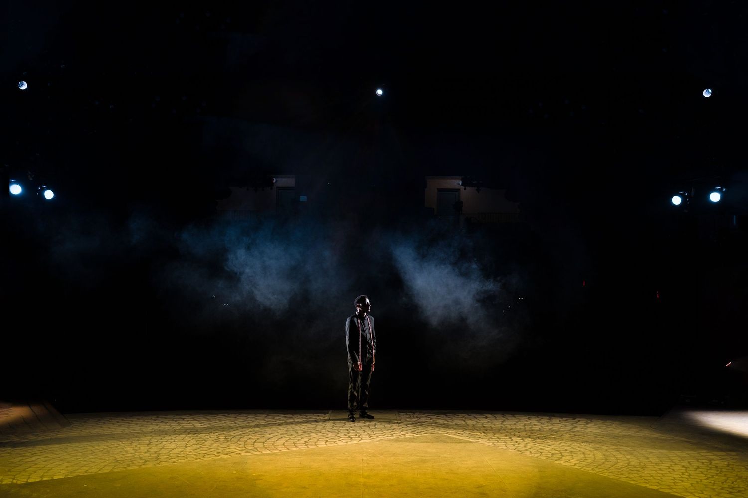 Lone performer stands in dramatic stage lighting with blue smoke and spotlights on a yellow-lit circular floor.