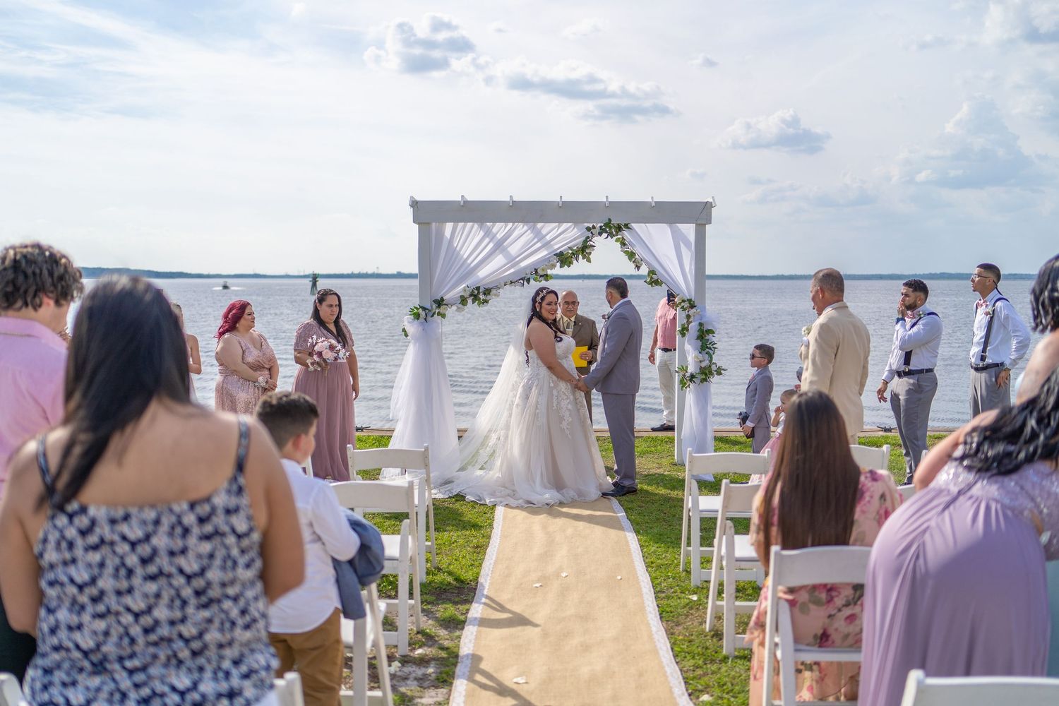 Outdoor waterfront wedding ceremony with bride and groom at floral arch, guests seated on white chairs under cloudy sky.