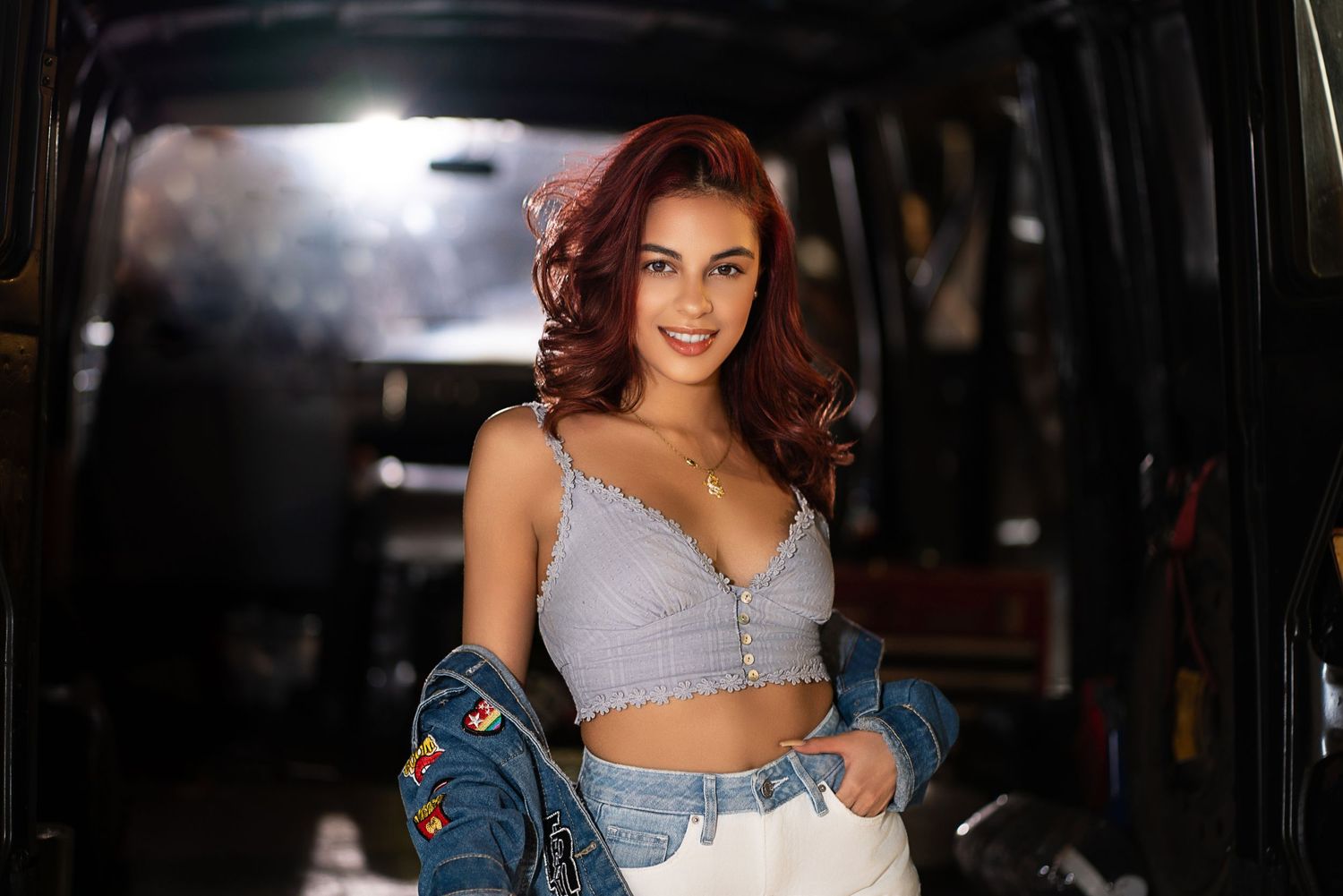 Smiling young woman with red hair wearing a crop top and denim jacket, posing in front of a van.